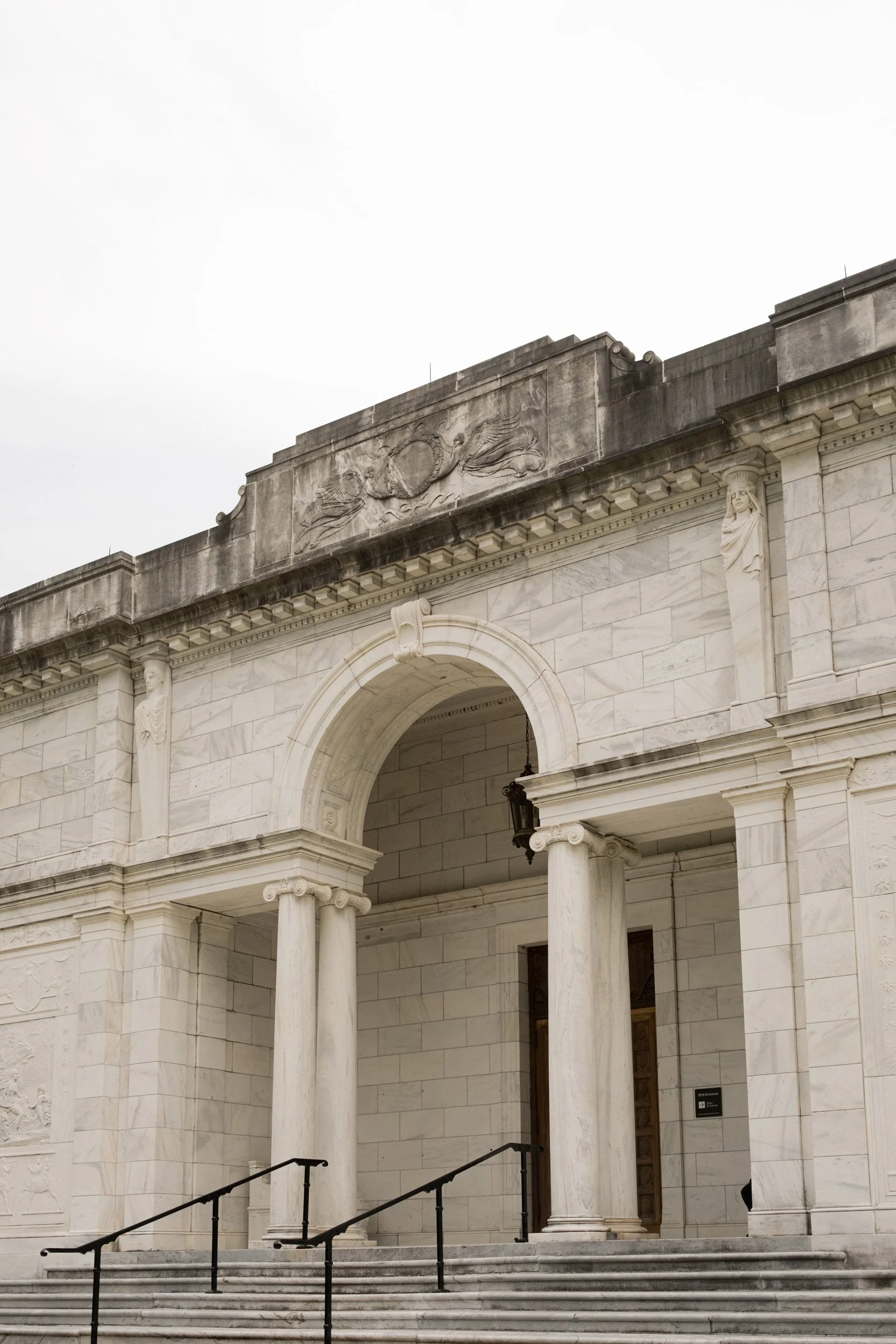 Stone building with columns, arched windows, and decorative architectural details, including a relief sculpture of a fish and a circular emblem, against a cloudy sky.