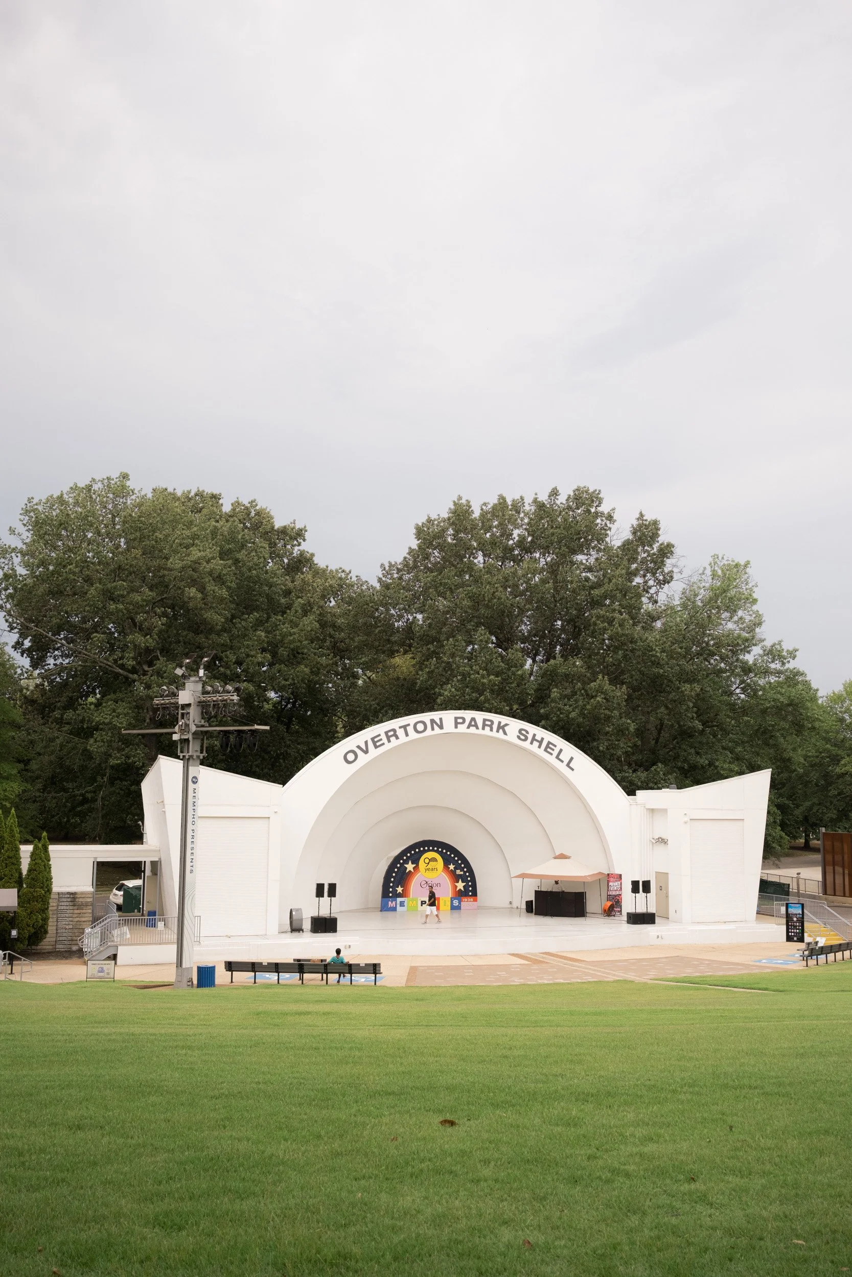 Empty outdoor stage with a white shell structure labeled "Overton Park Shell," set in a park with green grass, benches, and trees in the background under a cloudy sky.