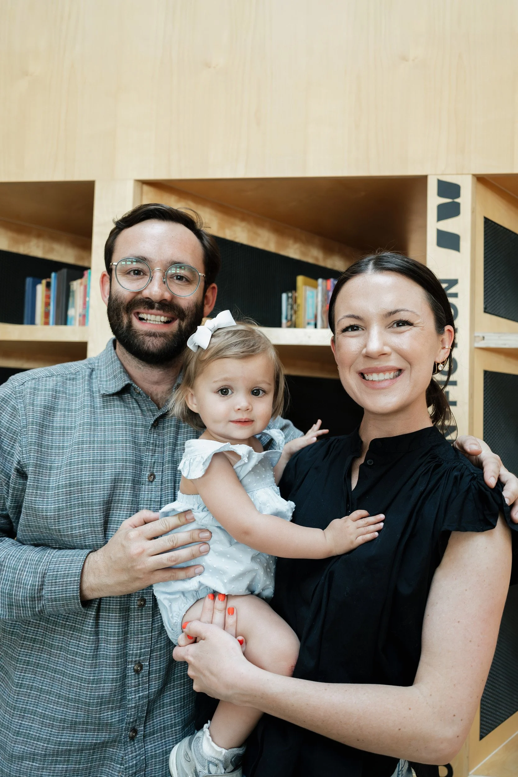 A happy family of four, including a man with glasses, a woman, and a young girl with a bow in her hair, standing in front of a bookshelf in a library or bookstore, smiling at the camera.