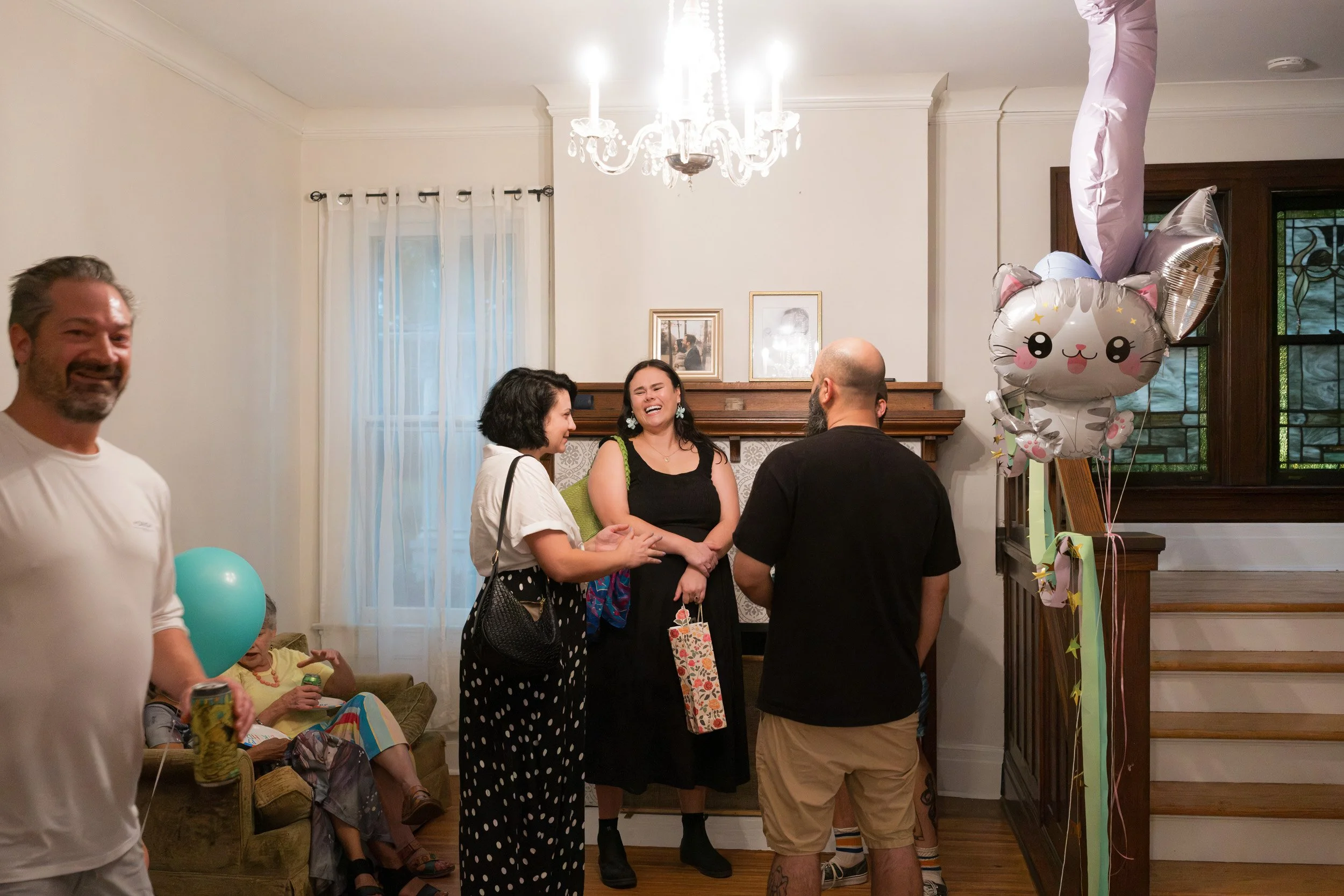 A group of people gathered in a living room during a celebration, with women laughing and talking, a man smiling in the foreground holding a can, and a baby sitting on a couch. There are birthday balloons, including a cute cat-shaped balloon, and a family photo on the wall by a fireplace.