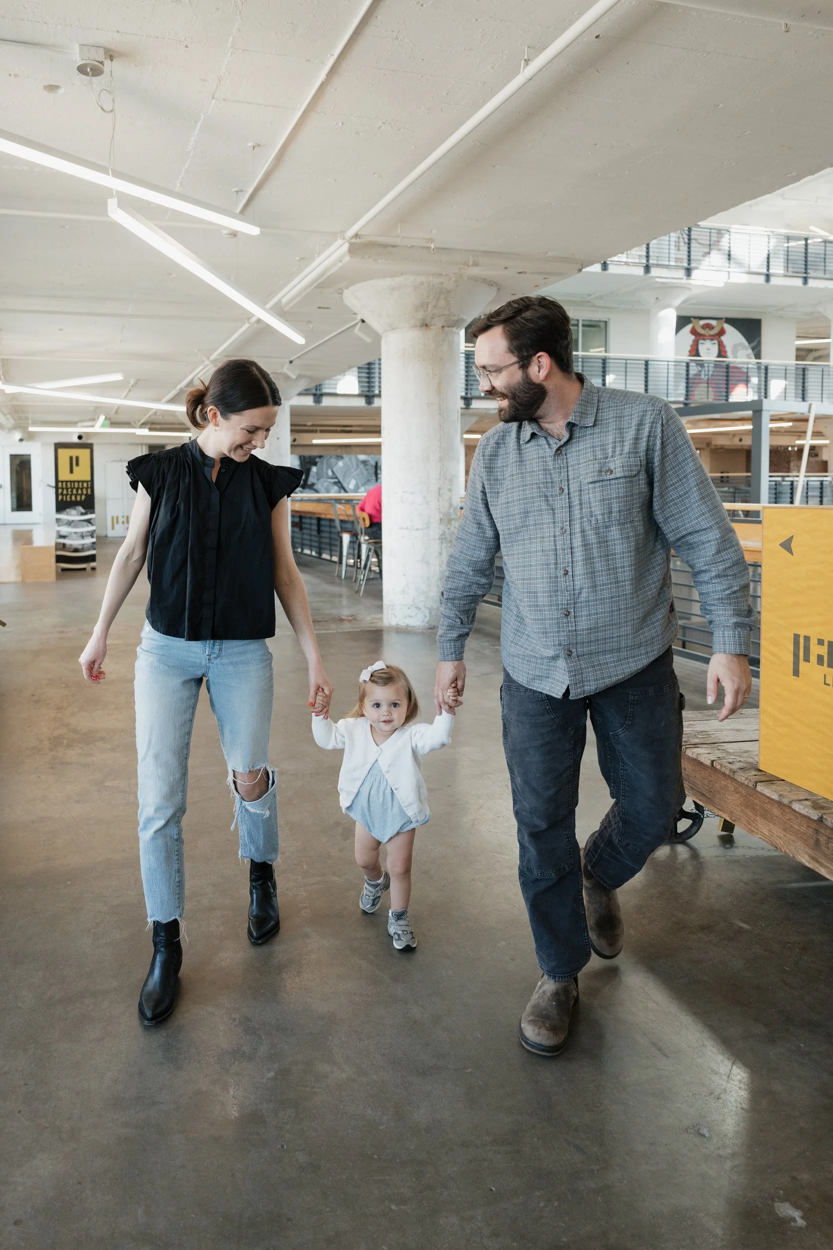 A family of four walking indoors at a modern building. The woman is wearing a black top, ripped jeans, and black boots. The man is wearing a gray plaid shirt and jeans. Their young daughter is holding hands with both, wearing a white jacket and blue dress.