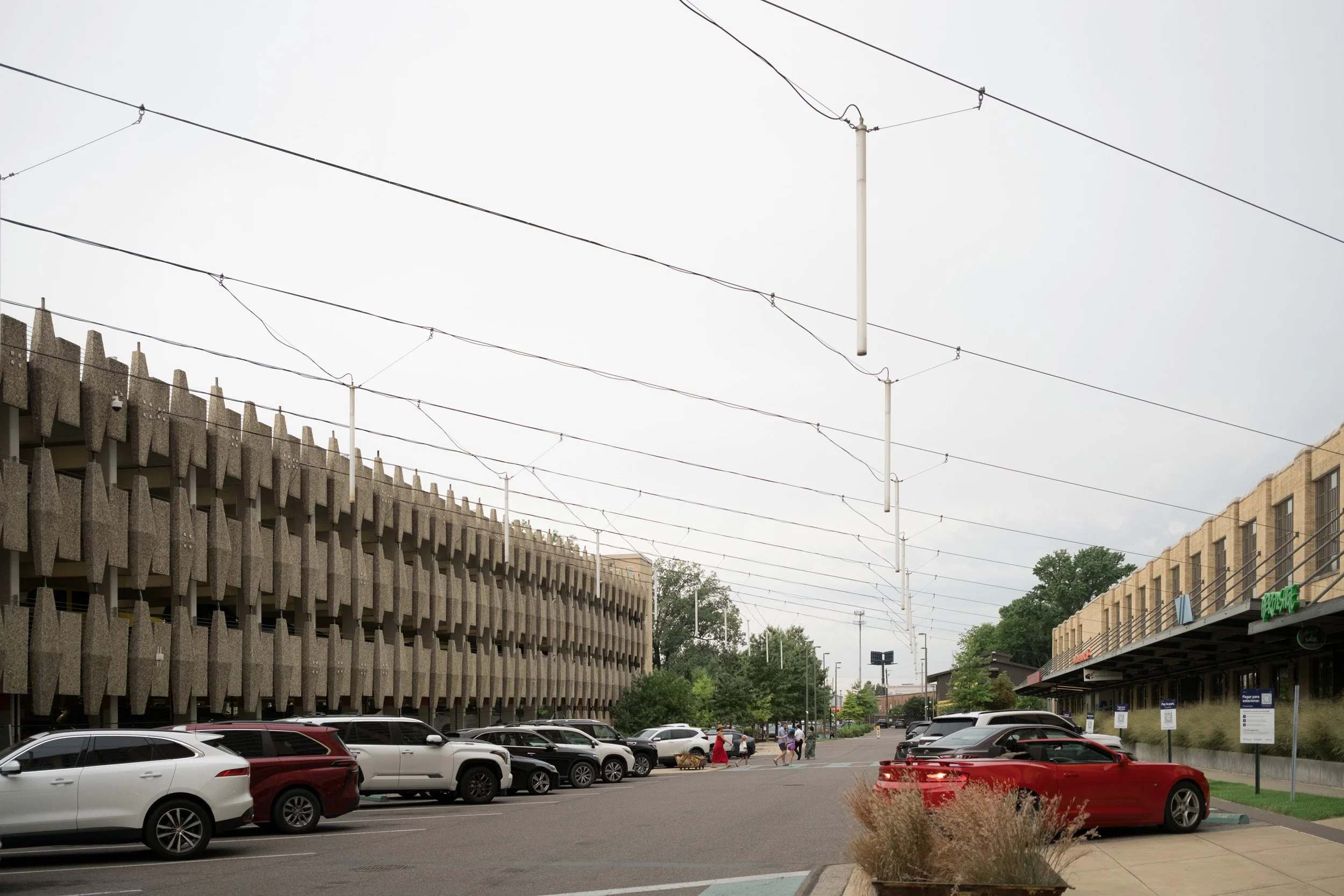 A parking lot with multiple cars parked and several people walking. There are buildings on either side, with the left building having a textured concrete facade and the right building appearing to be a commercial or retail space. Overhead, there are wires and poles, and the sky is overcast.