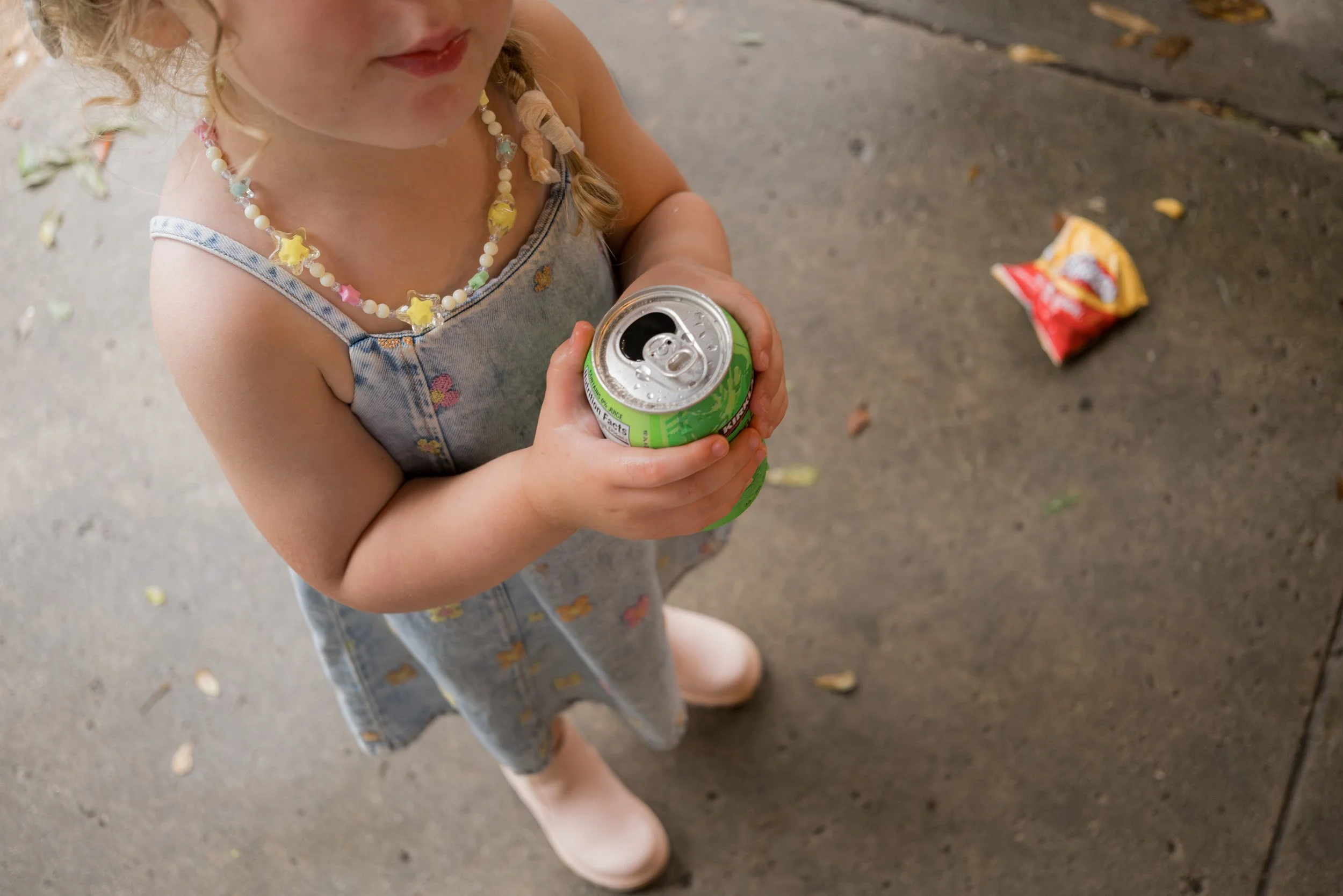 A young girl with curly blonde hair wearing a floral tank top, pastel-colored beaded necklace, and denim shorts, standing on a concrete sidewalk holding an open soda can. A crumpled snack bag is on the ground nearby.