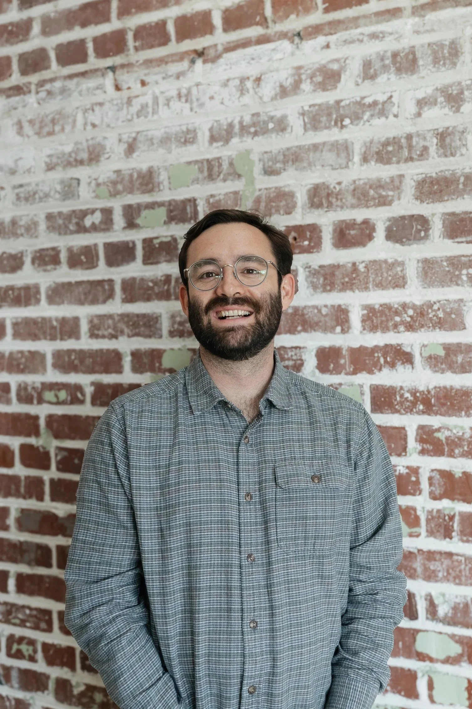A man with glasses and a beard standing in front of a red brick wall.