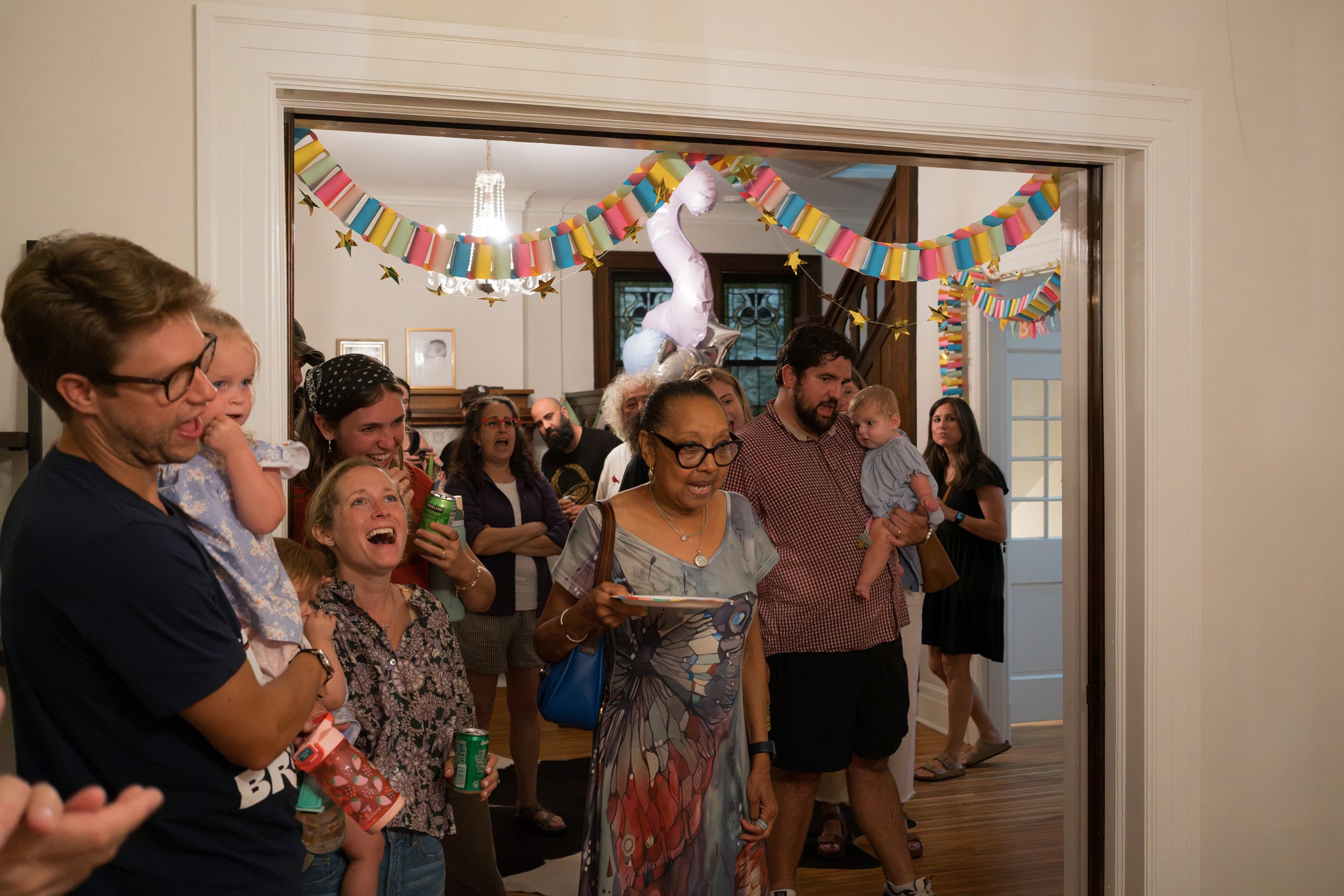 A group of people gathered in a room decorated for a celebration, with colorful paper garlands and balloons hanging from the ceiling.