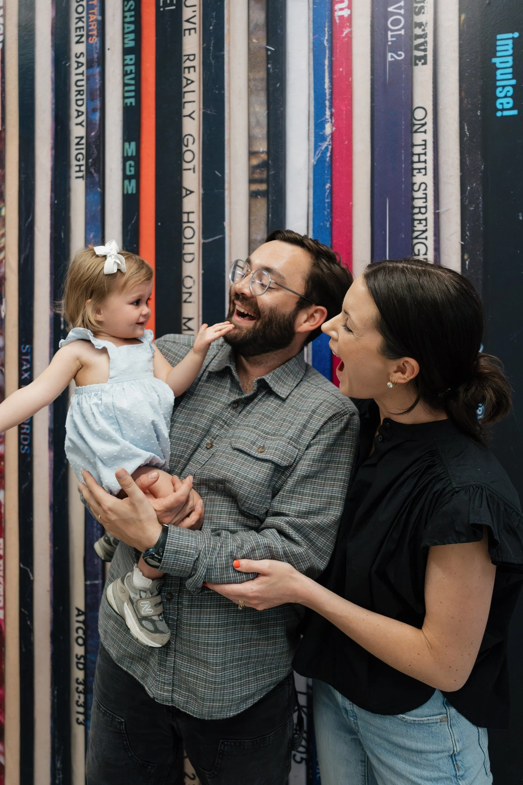 A man with glasses and a beard is holding a young girl in his arms, while a woman with dark hair and earrings is smiling at them. The girl, wearing a white dress with a white bow in her hair, is reaching out to touch the man's face. They are standing in front of a wall decorated with colorful vertical book spines.
