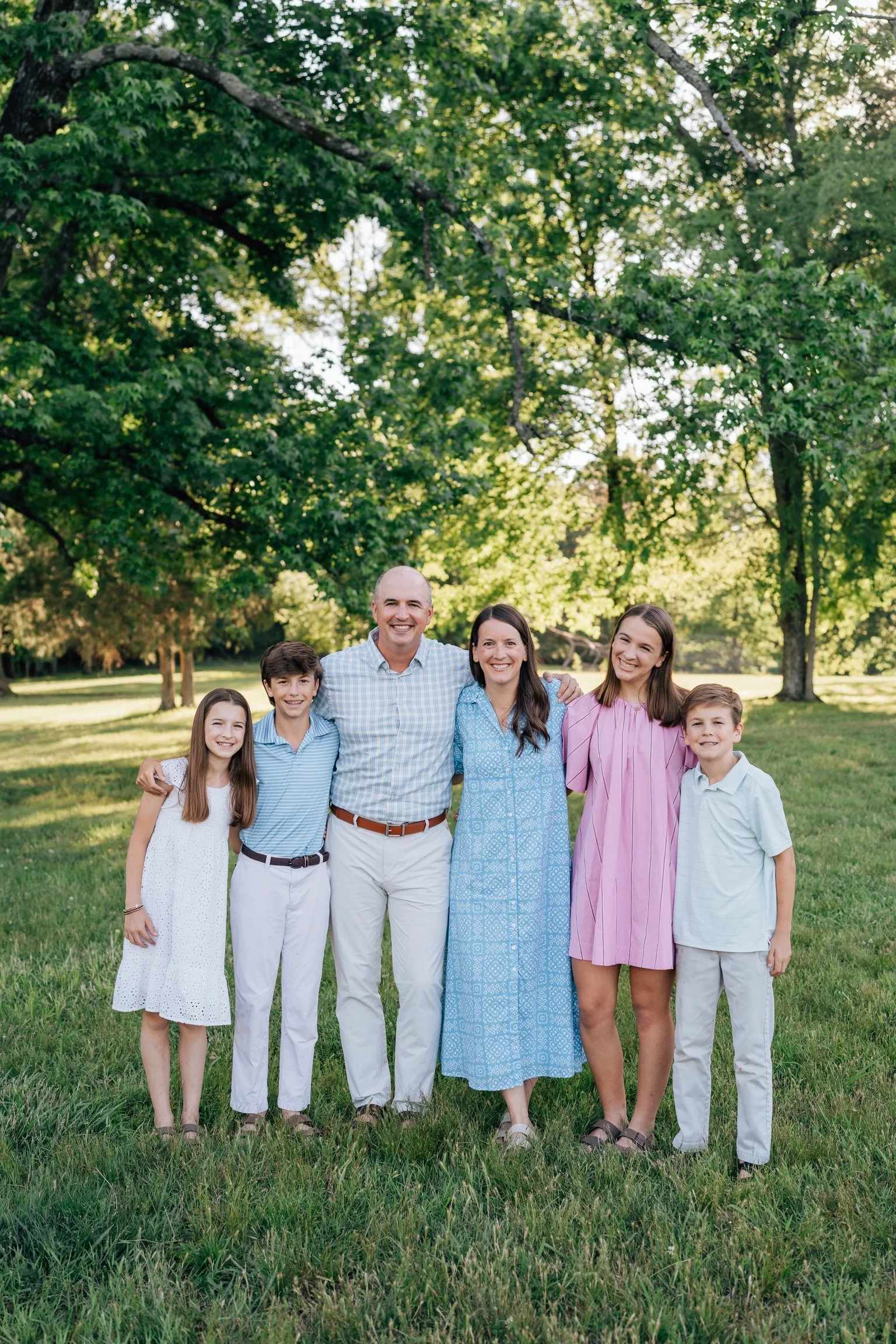 A family of six standing outdoors on a grassy field with trees in the background, smiling and posing for a photo.