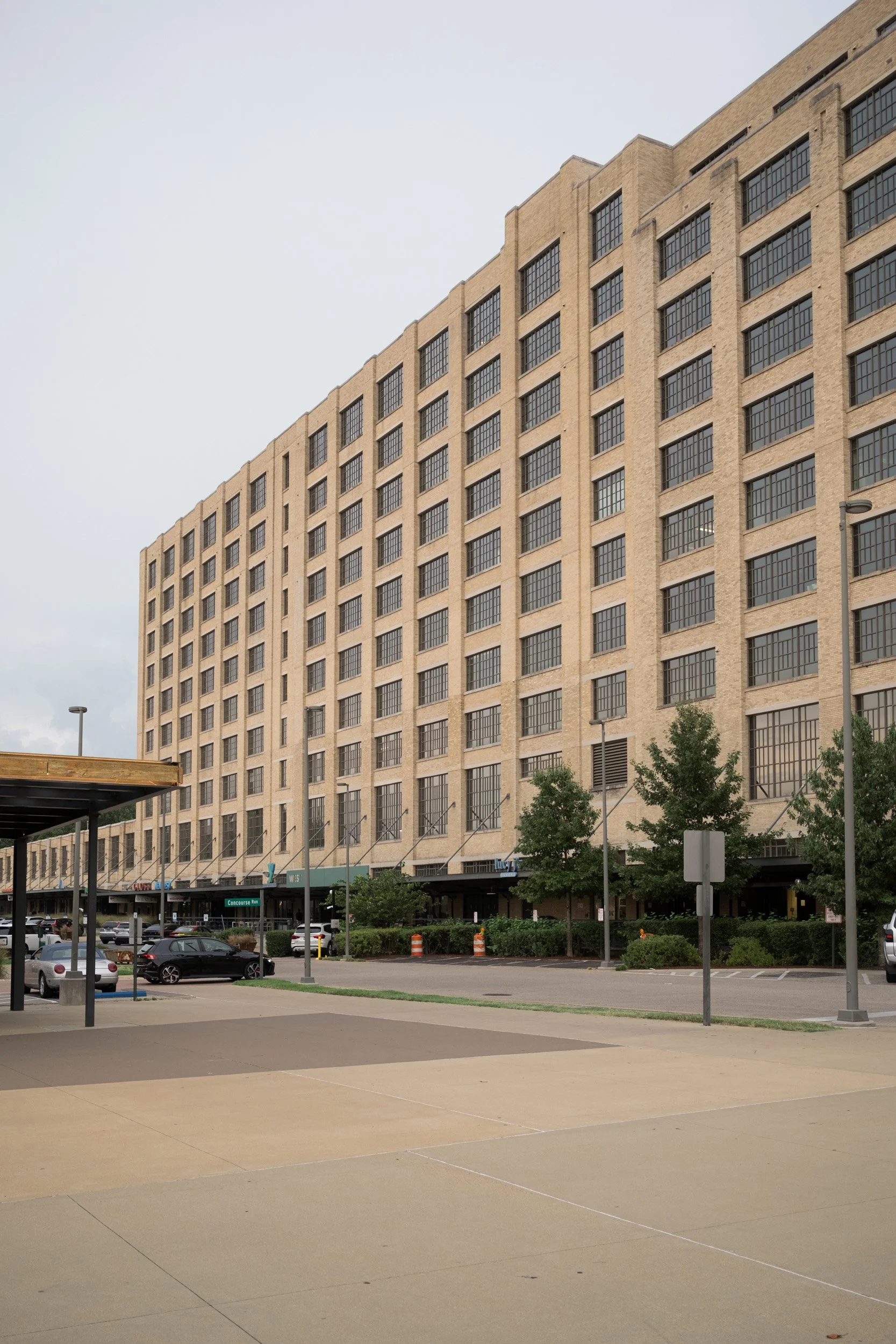 A large beige brick building with multiple rows of black-framed windows. In front, there is a parking lot with some cars, trees, and a small area of pavement.