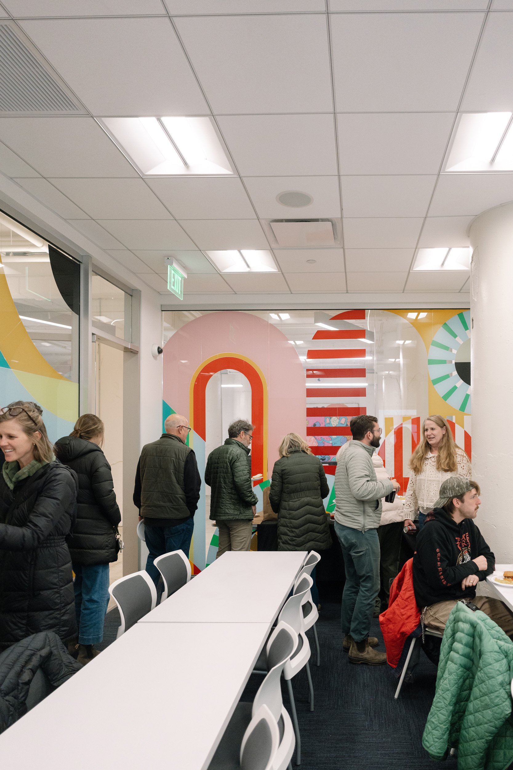 People are standing and sitting in a modern office or conference room with colorful wall designs. Some are talking, some are sitting at tables, and others are waiting in line.