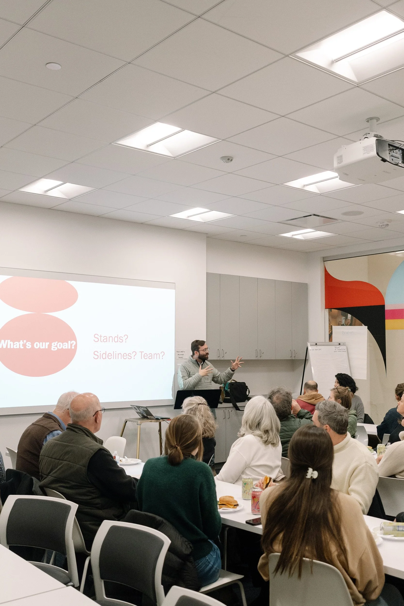 A man in a gray shirt giving a presentation to a group in a classroom or conference room setting. The audience is seated at tables with some people having food and drinks. There is a large screen displaying some questions and text related to a discussion or workshop. The room has a white ceiling with fluorescent lights and a projector mounted on the ceiling.