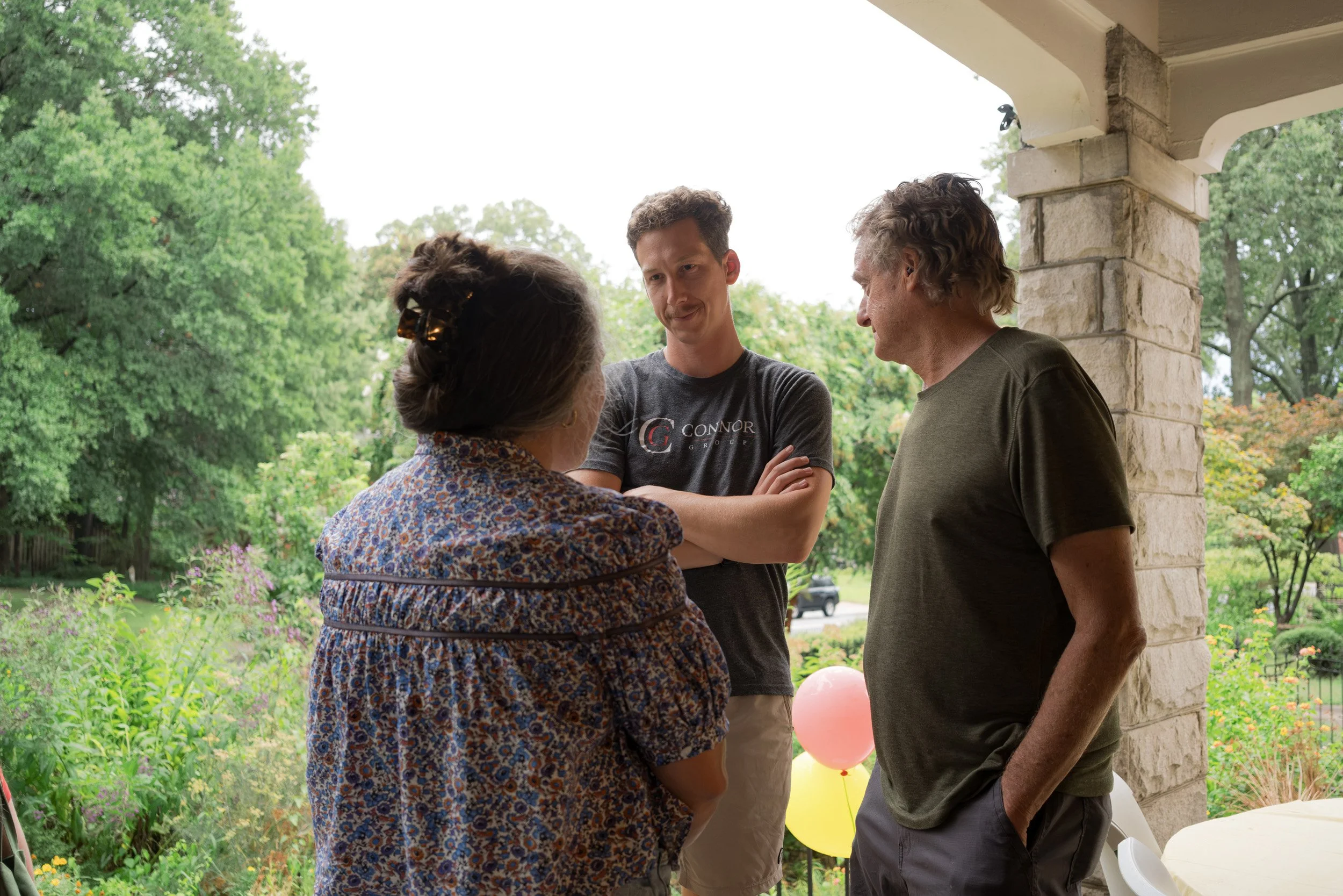 Four people standing on a porch engaged in conversation, with greenery and balloons in the background.