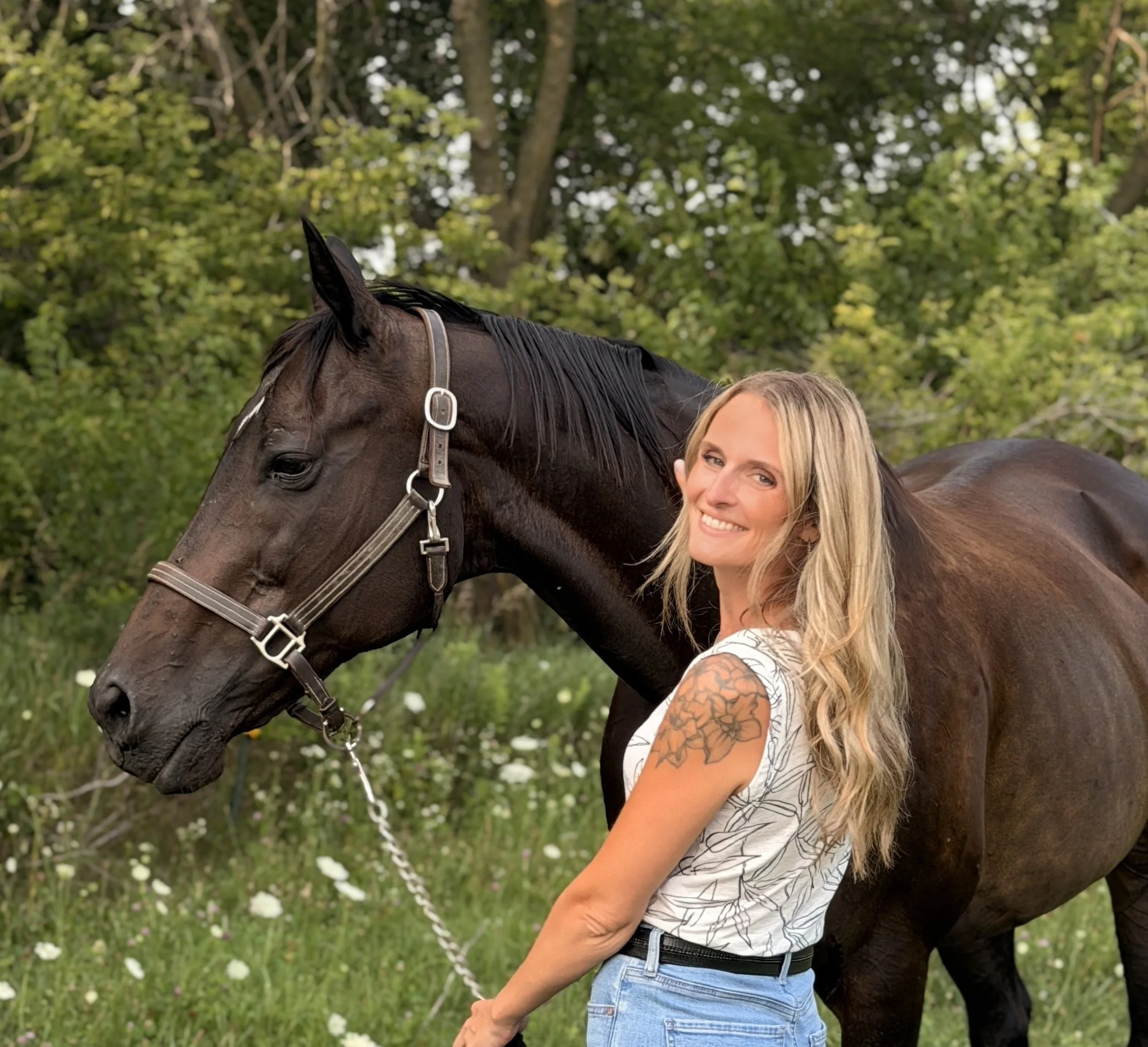 A woman with blonde hair and tattoos on her arm, smiling, standing next to a dark brown horse with a black mane outdoors on a grassy area with trees in the background.