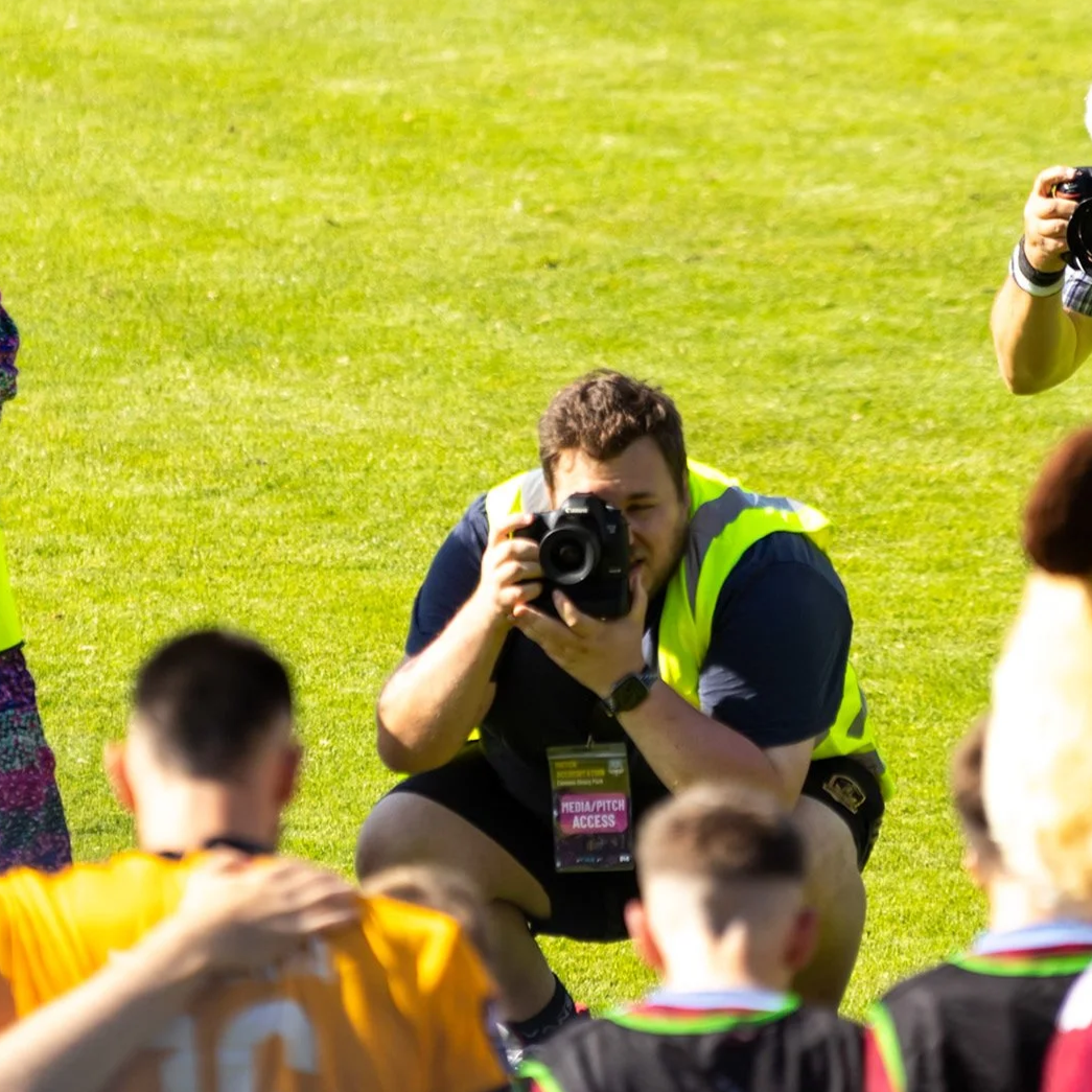 Photographer crouching on grass, holding a camera, taking pictures of players wearing sports jerseys during daytime outdoor event.