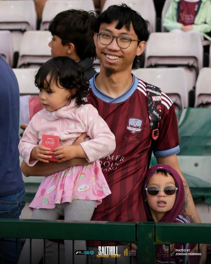 A smiling man with glasses and two children at a stadium or sports event, one girl with sunglasses and a hoodie, and a younger girl in a pink hoodie holding a red item.