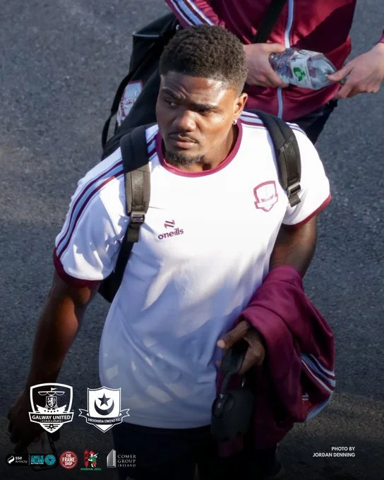 A young man with short hair wearing a white sports jersey with maroon accents, carrying a black backpack, holding a maroon jacket and a phone, standing outdoors on a dark paved surface, seen from above.