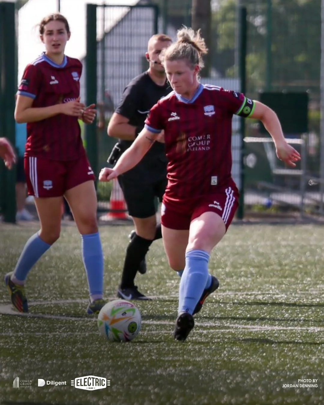 Female soccer players in maroon uniforms playing on a green field, with one about to kick the soccer ball while an official watches in the background.