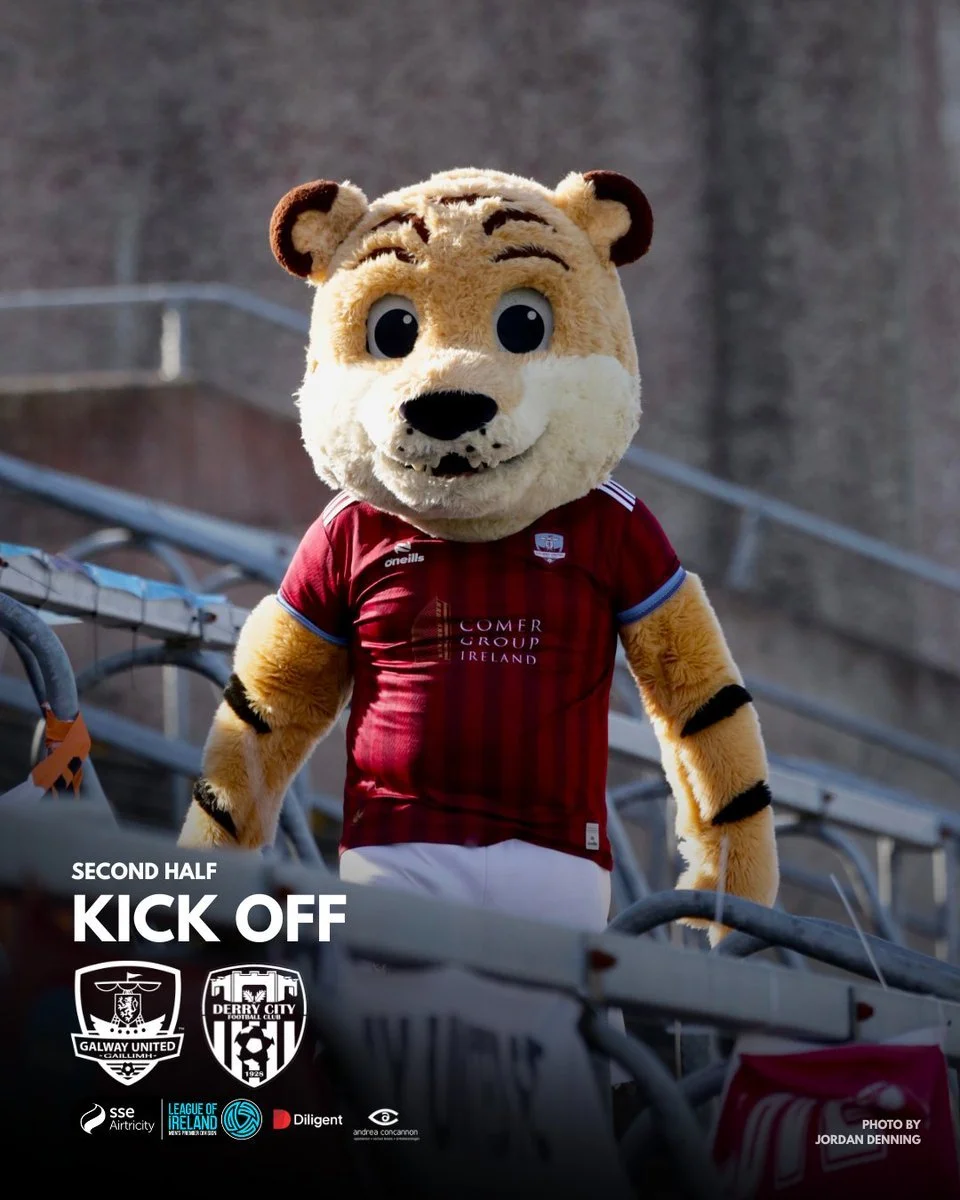 A person dressed in a tiger mascot costume wearing a red soccer jersey, standing in a stadium with empty bleachers.