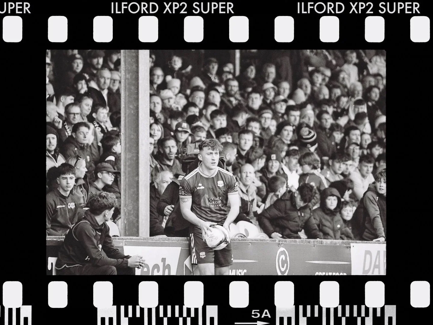 Black and white photo of a Soccer game with a young male player (Ed McCarthy) holding a  football, standing by the sideline. A large crowd of spectators is in the background, seated and standing, watching the game.