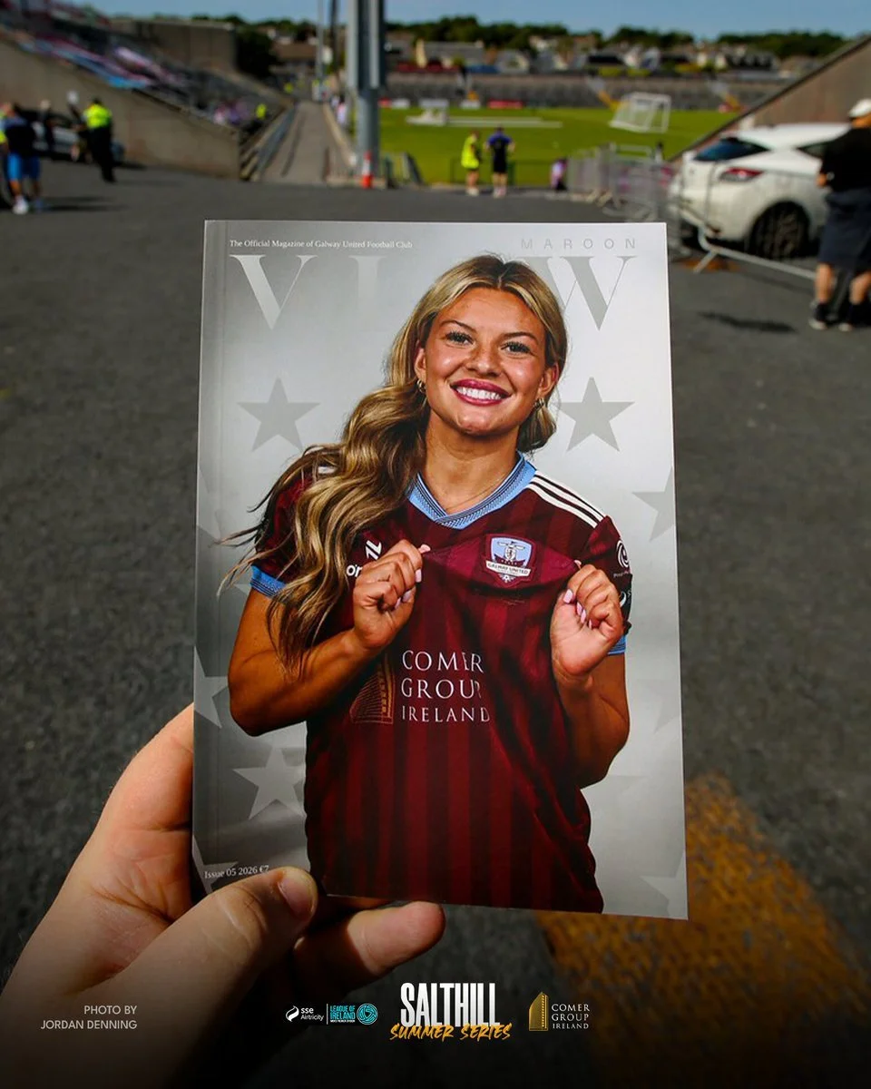 A person holding a magazine featuring a smiling woman in a maroon sports jersey with her fists raised. The background shows a sports stadium with fans, cars, and a grass field.