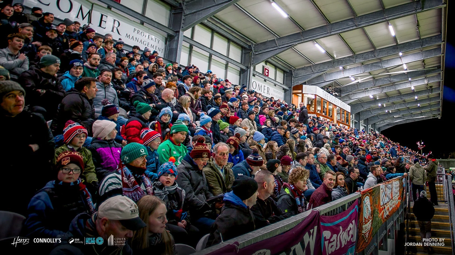 Crowd of spectators sitting in stadium stands, many wearing winter hats and jackets, at a sporting event at night.