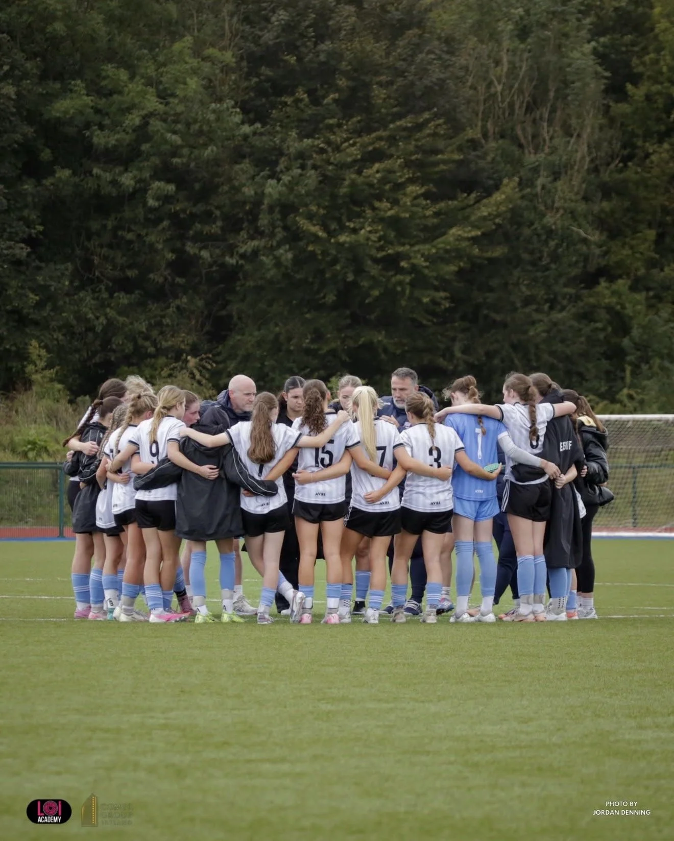 A group of female soccer players in white jerseys and black shorts, with some wearing light blue socks, stand together on the field with their arms around each other, listening to their coaches during a team huddle on a soccer field with a green and 