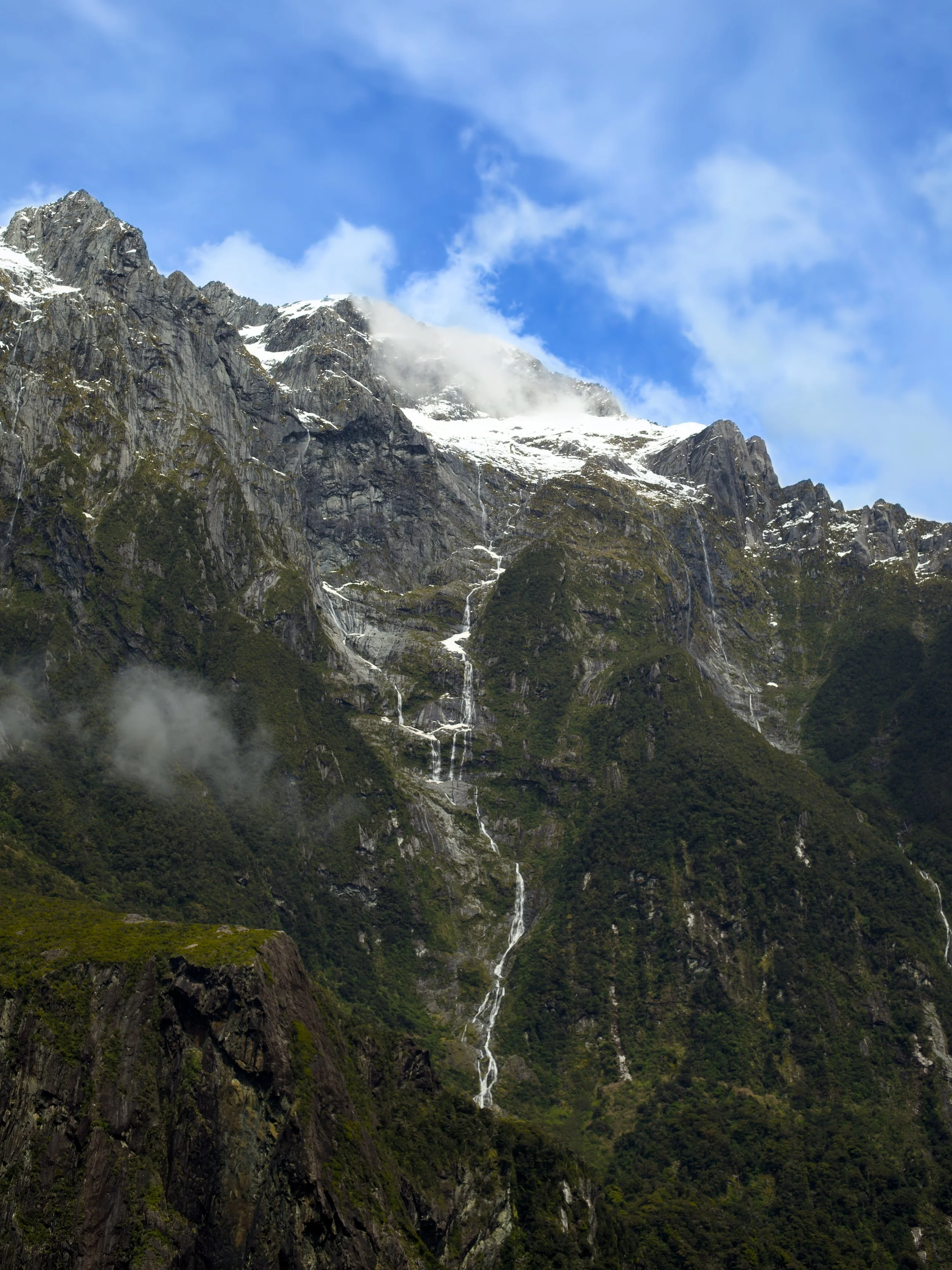 Mountain cliff with snow patches, waterfalls flowing down, and lush green forest at the base, under a partly cloudy blue sky.