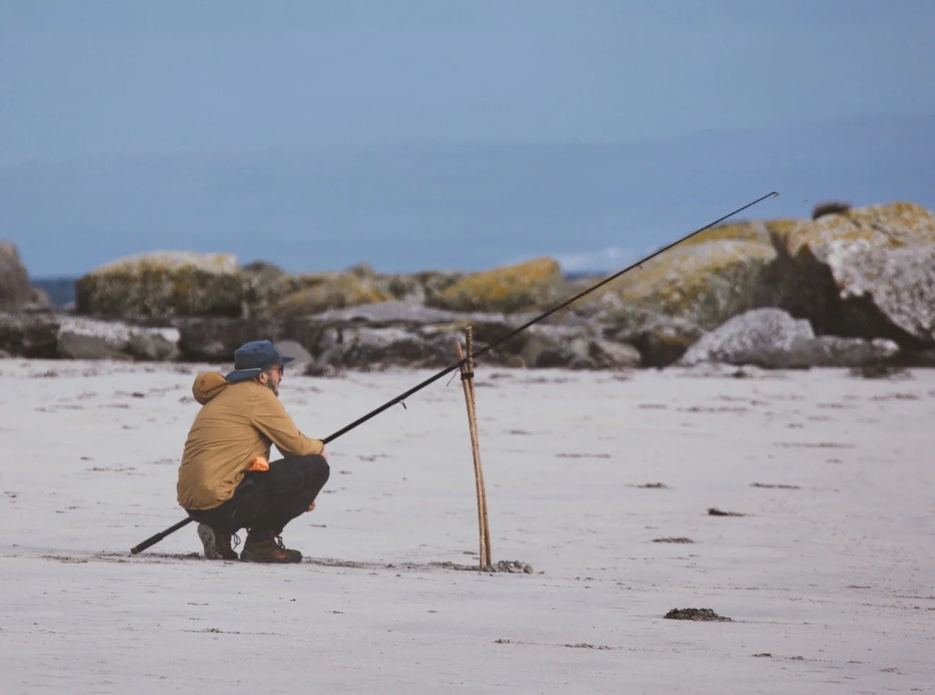 A man squatting on a sandy beach, holding a fishing pole, with rocks in the background and the ocean in the distance.