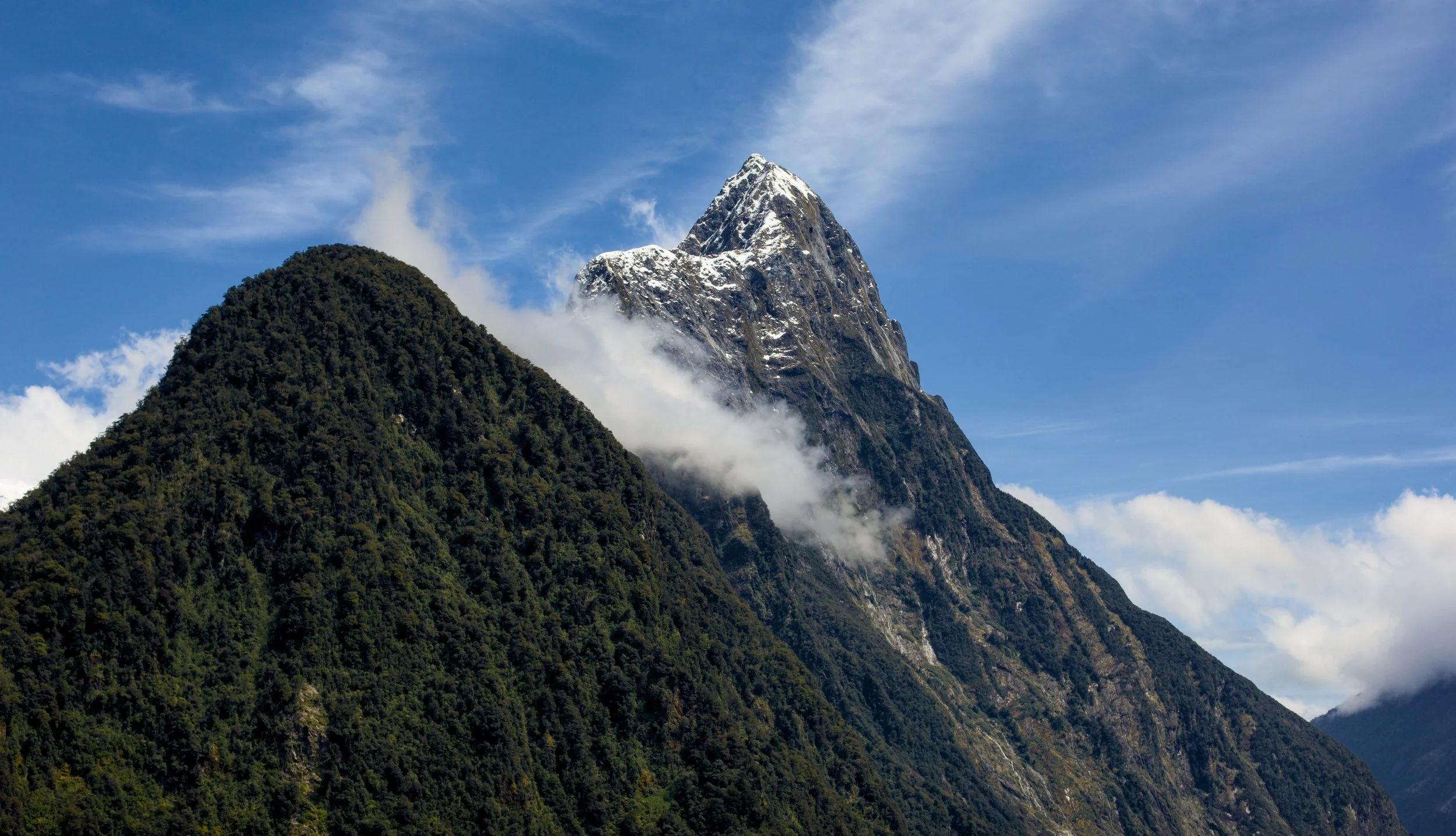 Snow-capped mountain peak rising above green forested hills with clouds and blue sky in the background.