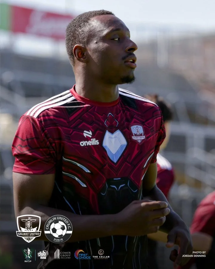 A young male soccer player wearing a red and black jersey with chameleon-inspired design, standing outdoors on a sunny day with stadium seating in the background.
