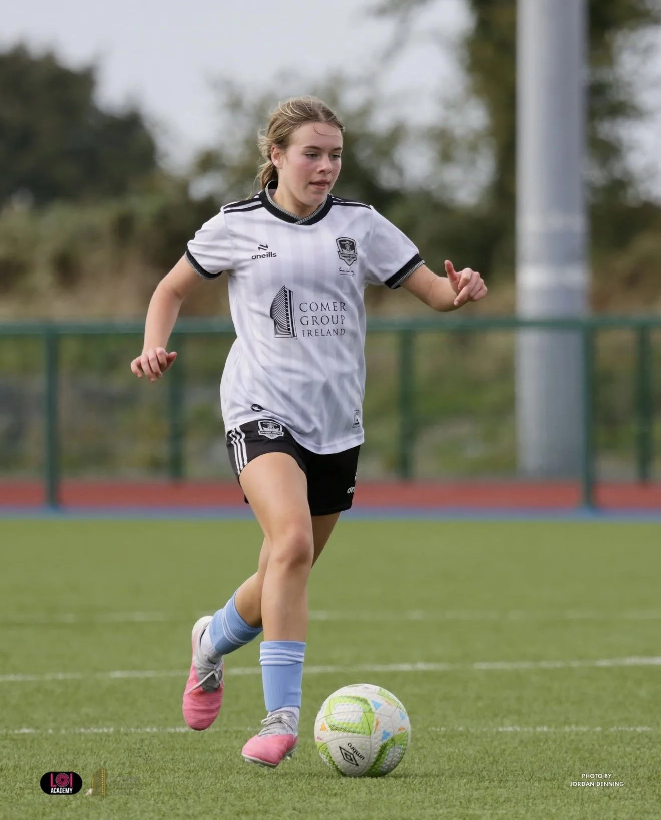 A young female soccer player in a white jersey and black shorts runs on a soccer field, dribbling a soccer ball with her right foot. She is wearing pink cleats and light blue socks, and her hair is tied back. In the background, there is a green fence