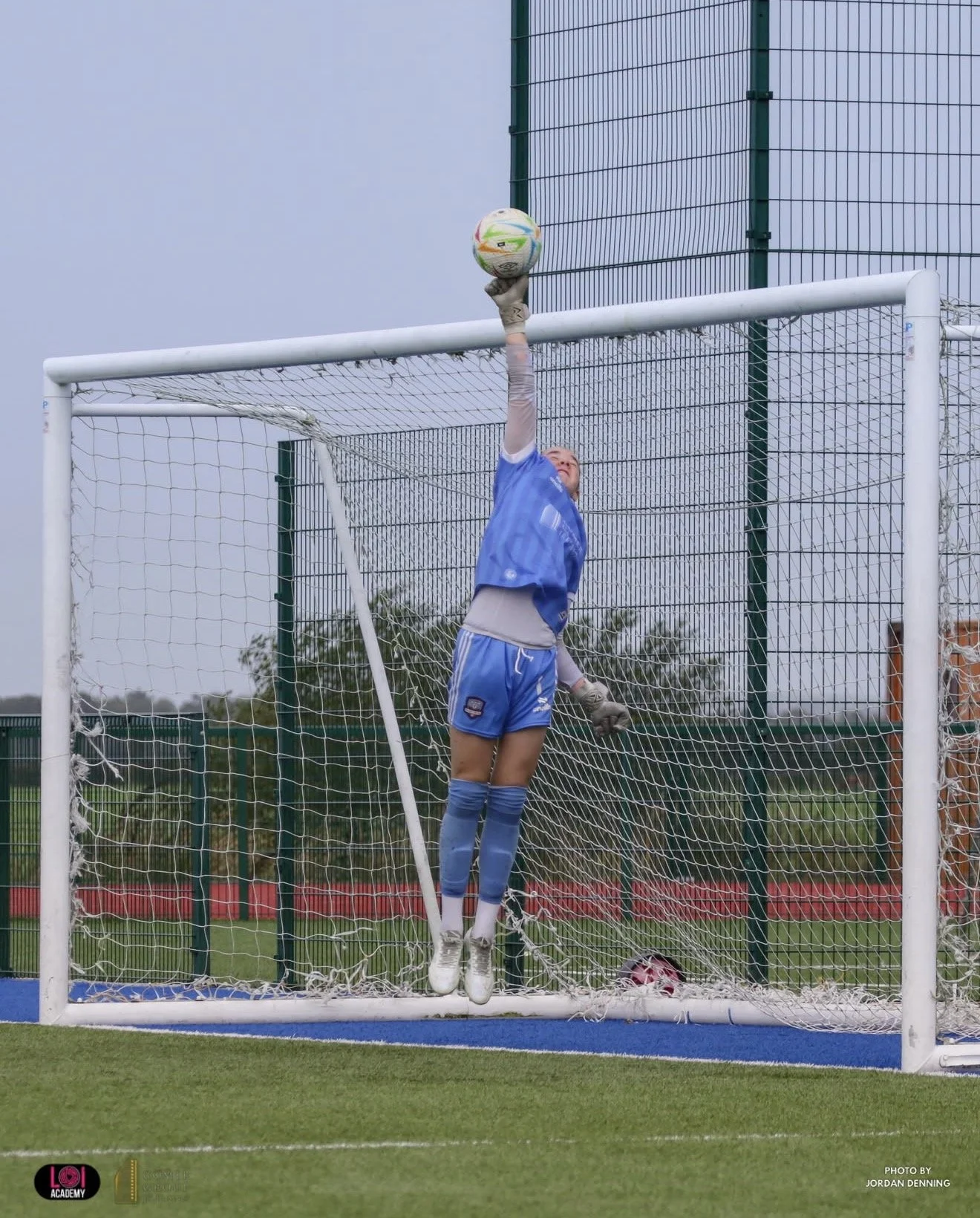 A female soccer goalkeeper in a blue uniform and gray gloves jumping and reaching up to catch a soccer ball inside the goal on a soccer field.