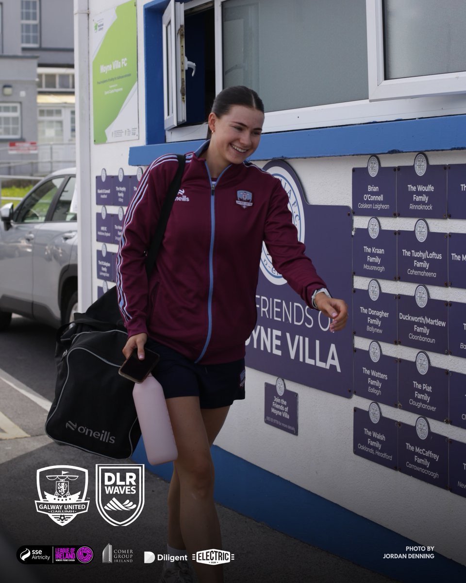 A young woman in sports attire smiling and walking past a community wall with her sports bag and water bottle, in front of a building with logos for Galway United and DLR Waves soccer teams.
