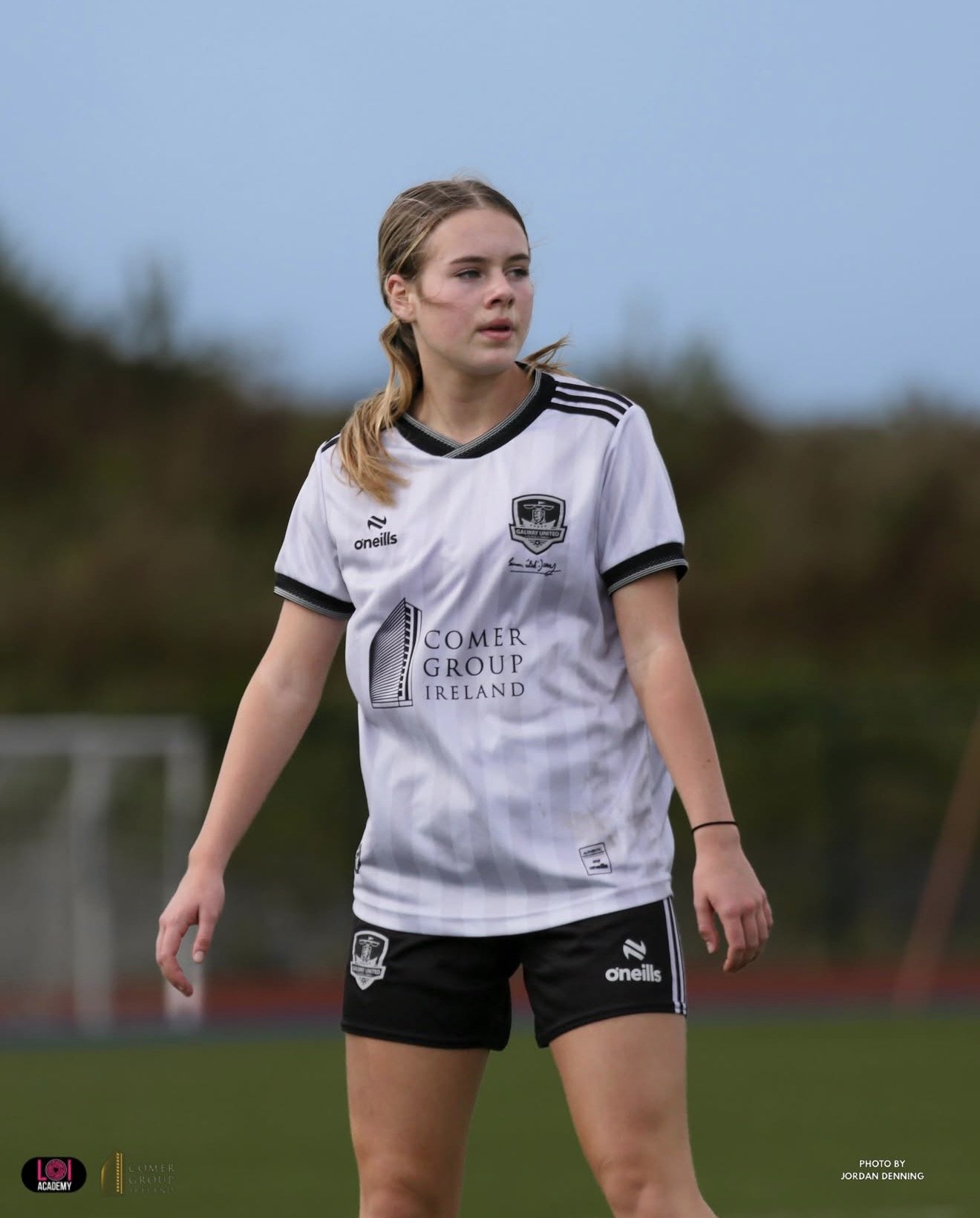 A young female soccer player in a white and black uniform on a field, with a cloudy sky in the background.