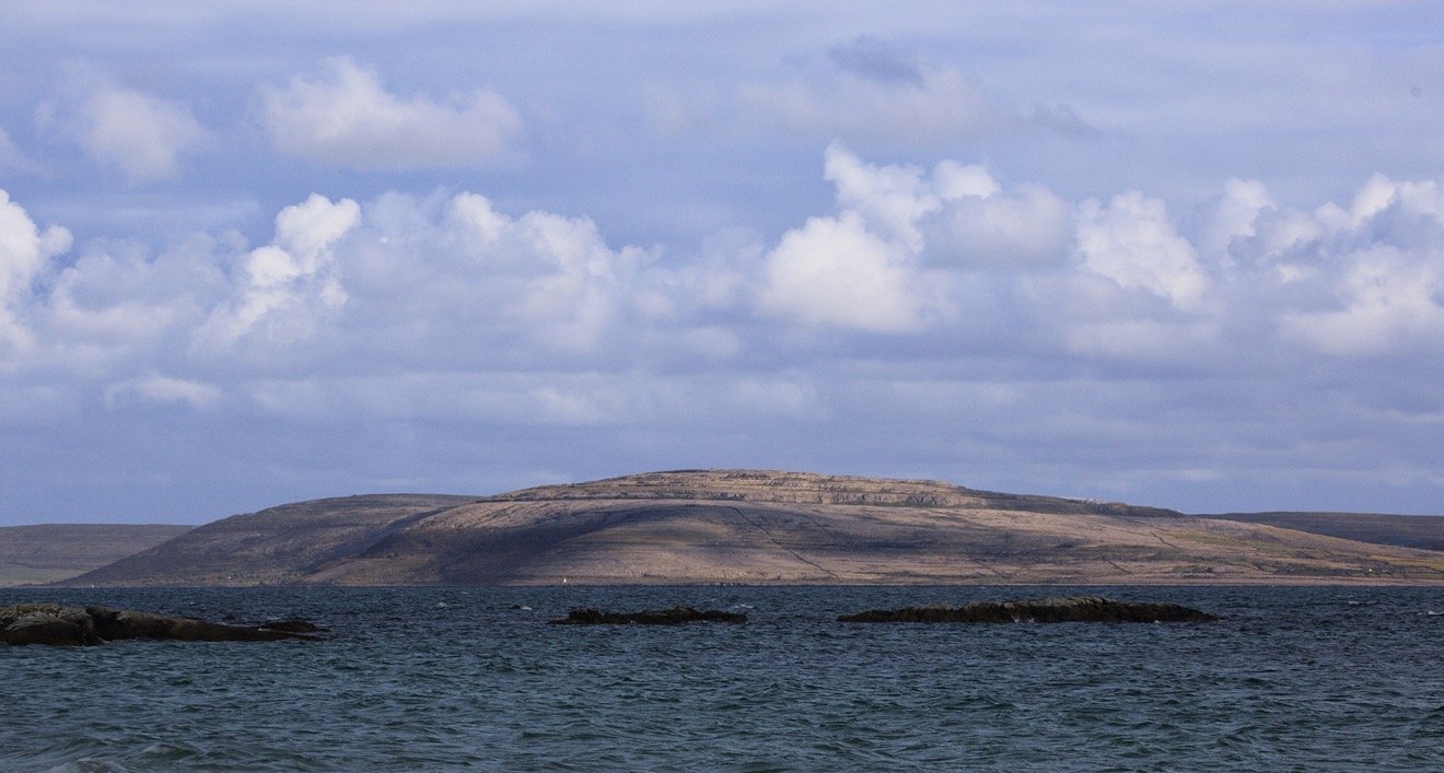A body of water with rocks in the foreground and a flat-topped hill in the background under a cloudy sky.