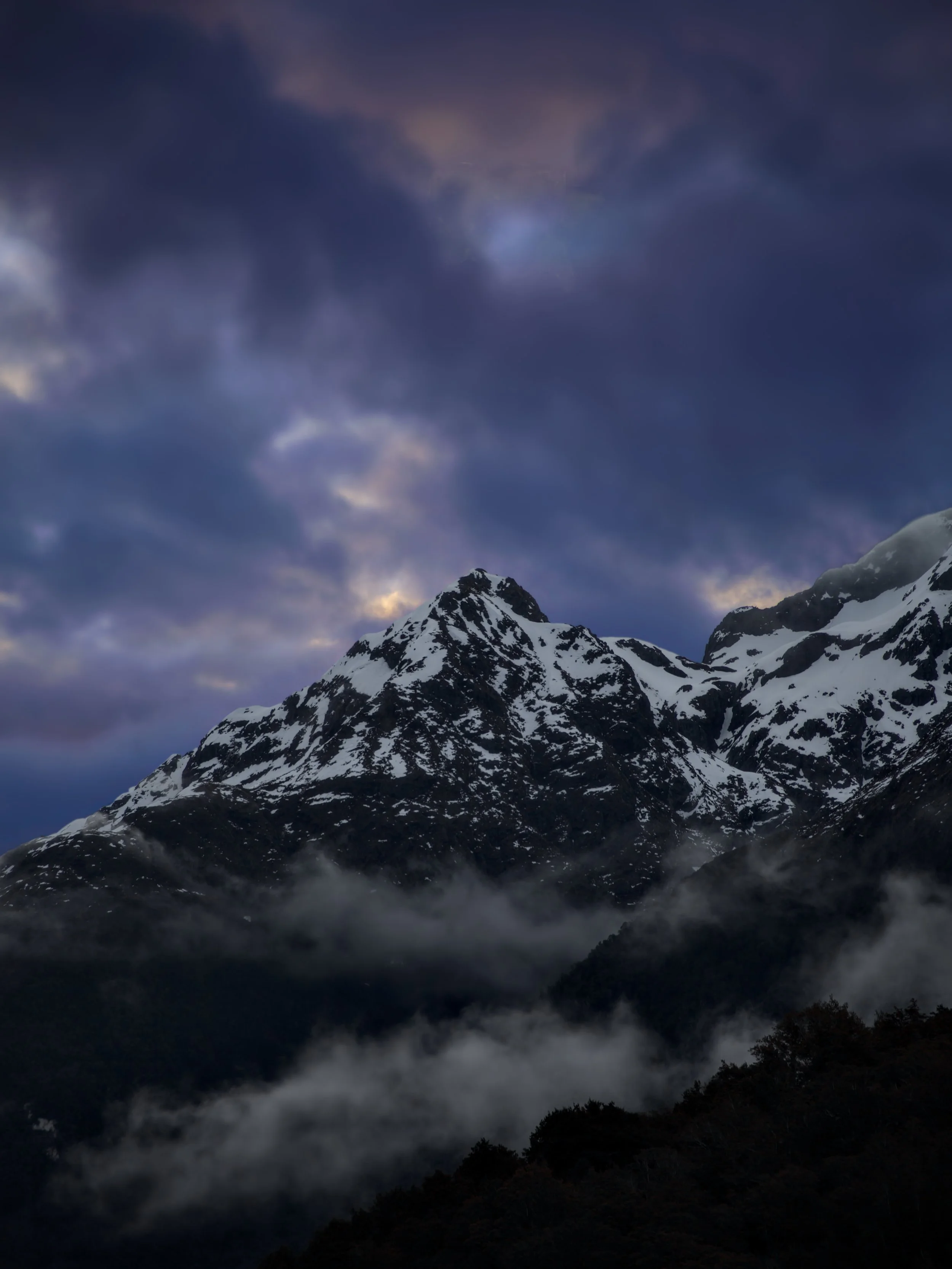 Snow-capped mountain peaks under a cloudy sky with patches of light peeking through, with mist or fog in the lower area.