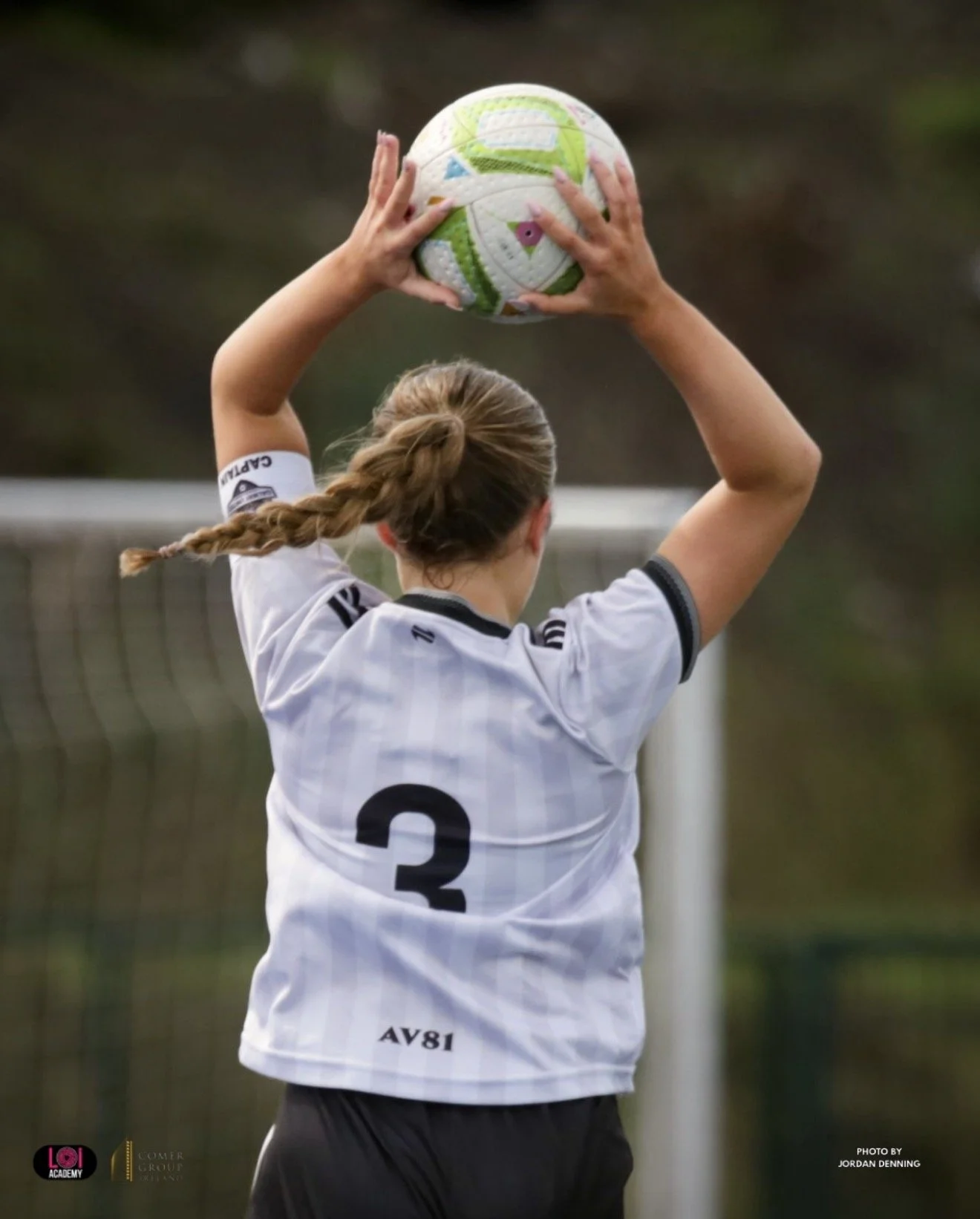 A female soccer player with braids, wearing a white jersey with the number 3, is holding a soccer ball above her head on the field.