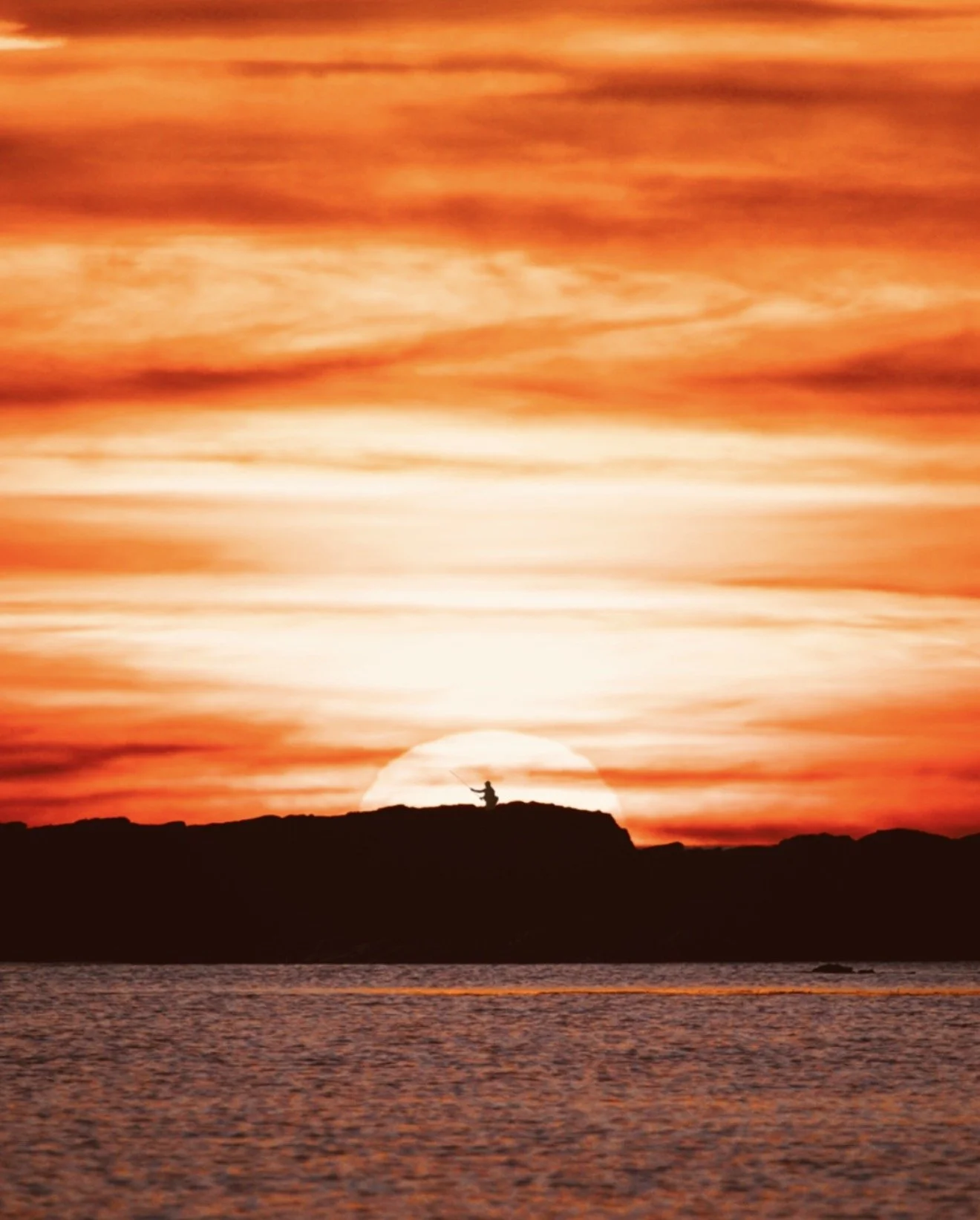 Silhouette of a person sitting on a rock facing the ocean during a vibrant sunset with a large sun near the horizon, colorful sky with orange and red clouds, and calm water in the foreground.