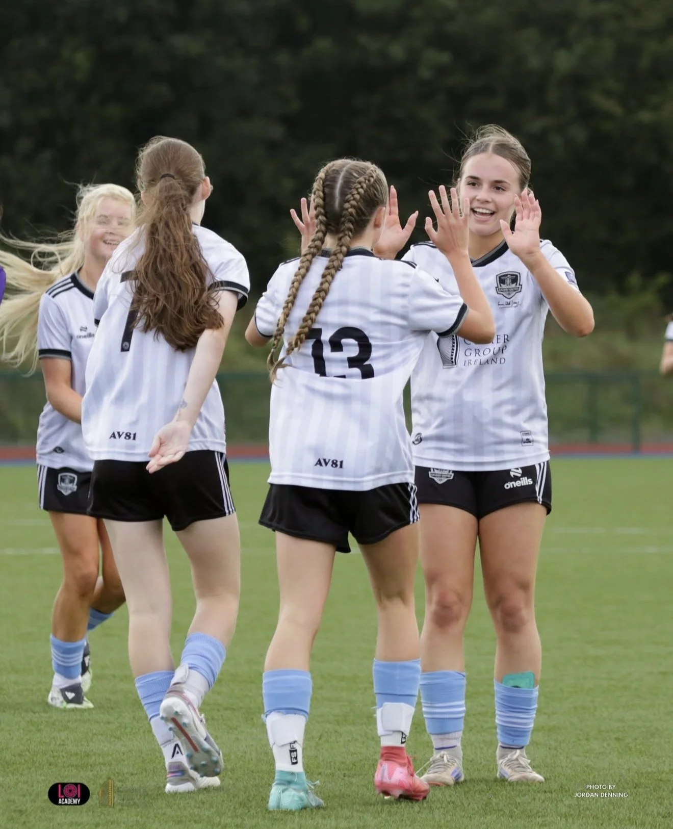 Group of young female soccer players celebrating on the field, wearing white and black uniforms and blue socks.