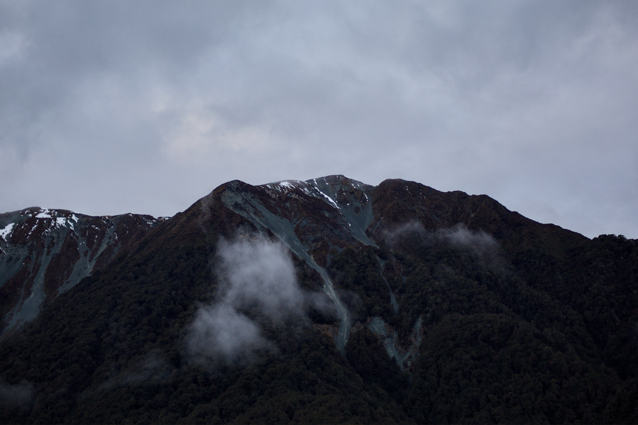 Mountain range with snow patches, dark rocky slopes, cloudy sky, and mist or smoke at the base.