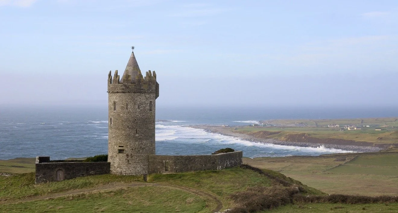 A medieval stone tower on a grassy hill overlooking the ocean, with a distant coastline and a cloudy sky.