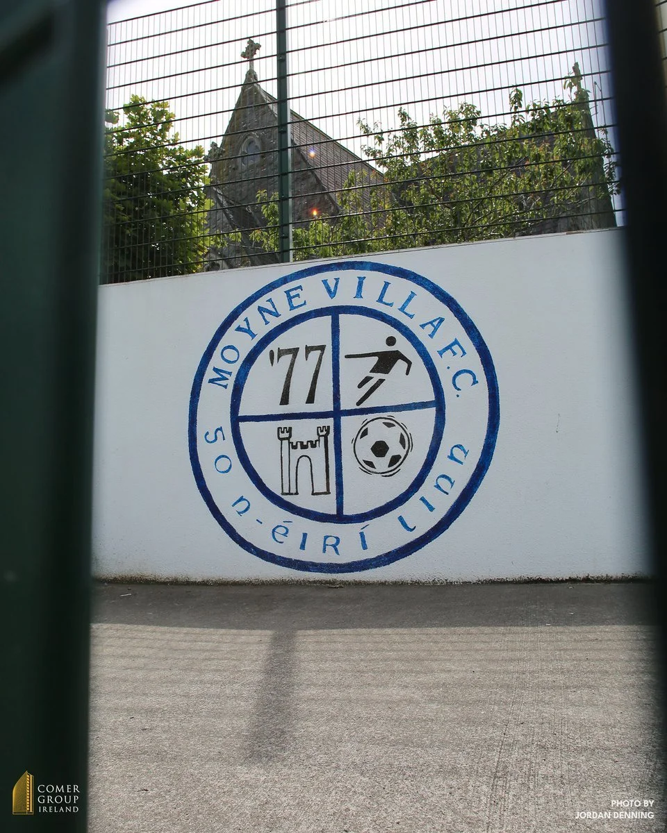 Fenced wall with Moone Village Football Club logo featuring a castle tower, soccer ball, running figure, and the year '77', with a church steeple and trees visible behind the fence.