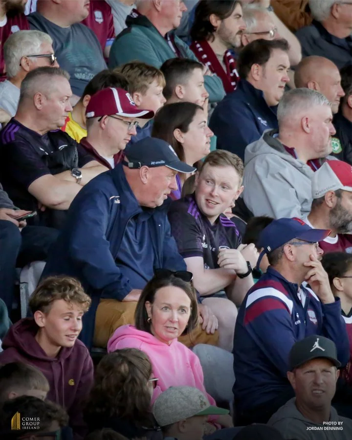 Crowd of sports spectators sitting in stadium seats, wearing jackets and caps, engaged in watching an event, with some talking and others focused ahead.