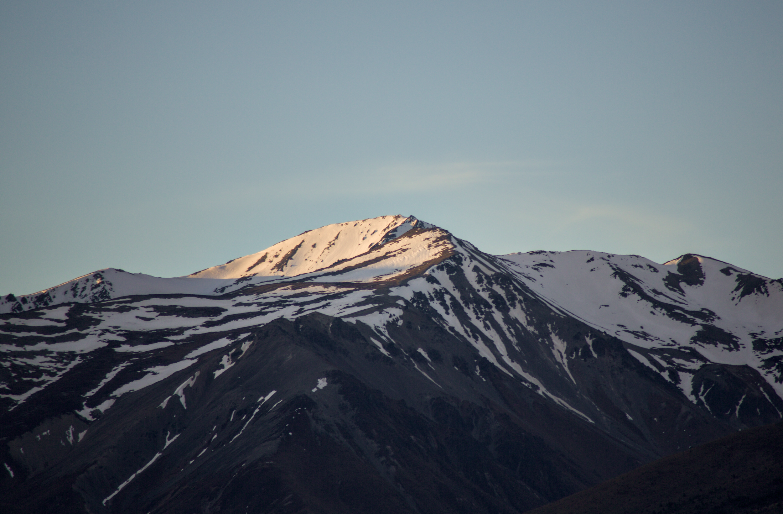 Snow-capped mountain peaks with patches of dark rock and snow, under a pale blue sky.