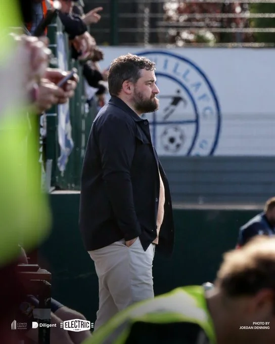 A man with a beard and short hair standing on a sports field sidelines, wearing a dark jacket and light-colored pants, with hands in pockets, watching a game.