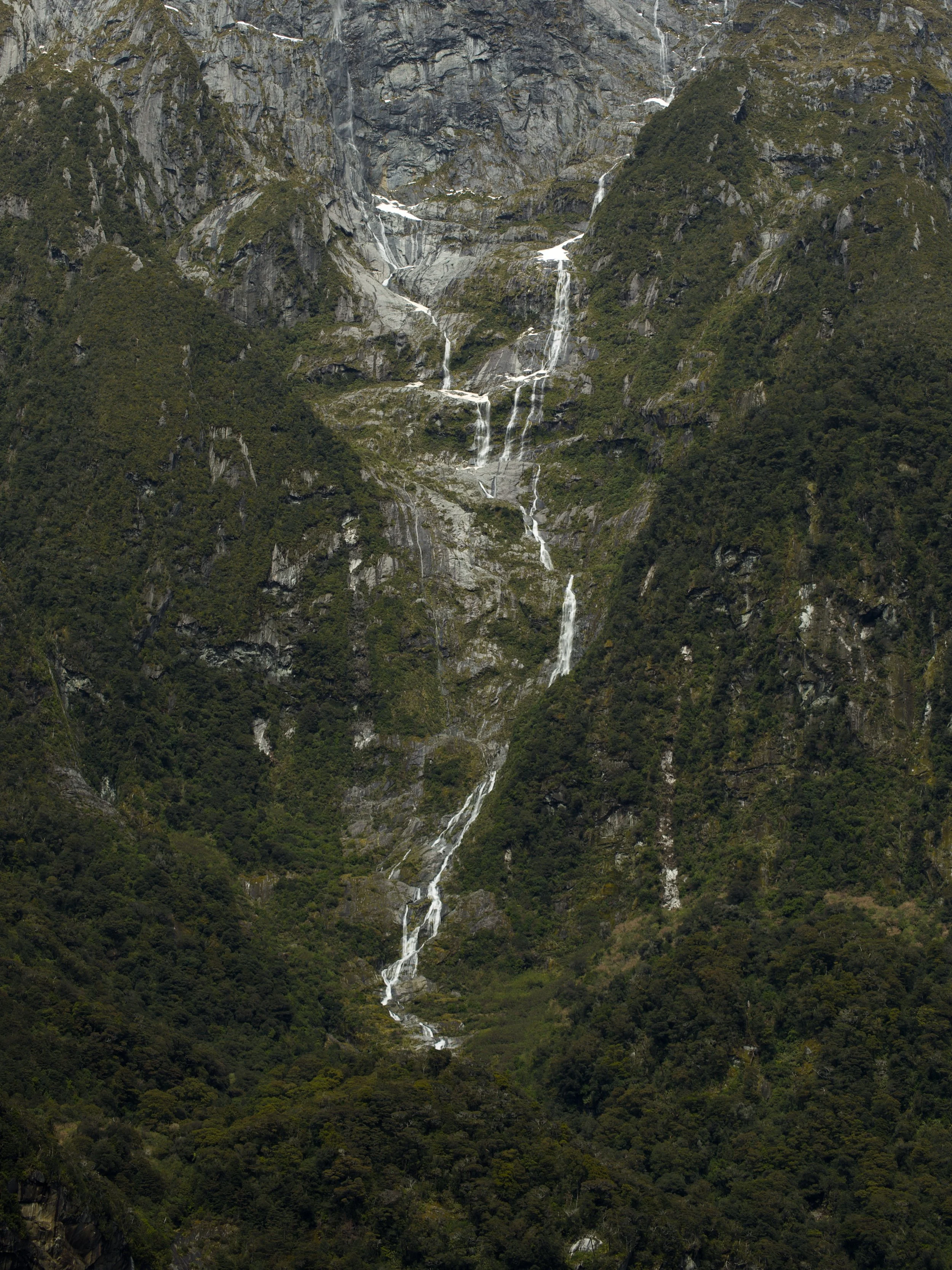 A mountain with steep, rocky cliffs and several thin waterfalls flowing down its face, surrounded by lush green trees.