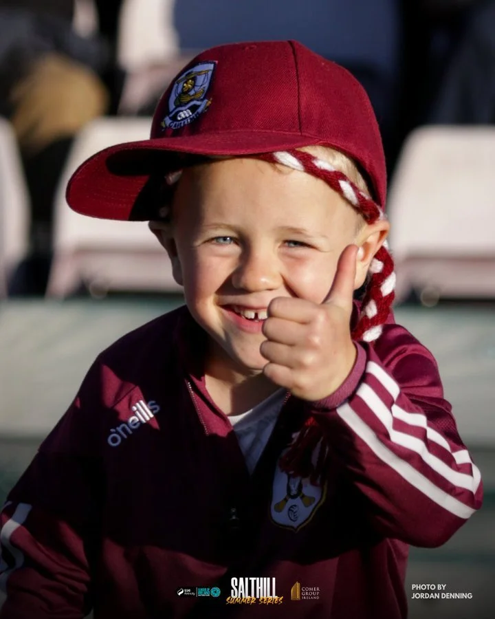 A young boy wearing a maroon sports jacket and a maroon cap with a sports team logo, smiling and giving a thumbs up, outdoors on a sunny day.