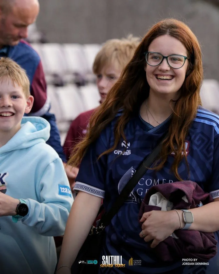 Smiling young woman with long red hair and glasses, wearing a blue sports jersey, standing among children at a sports event.