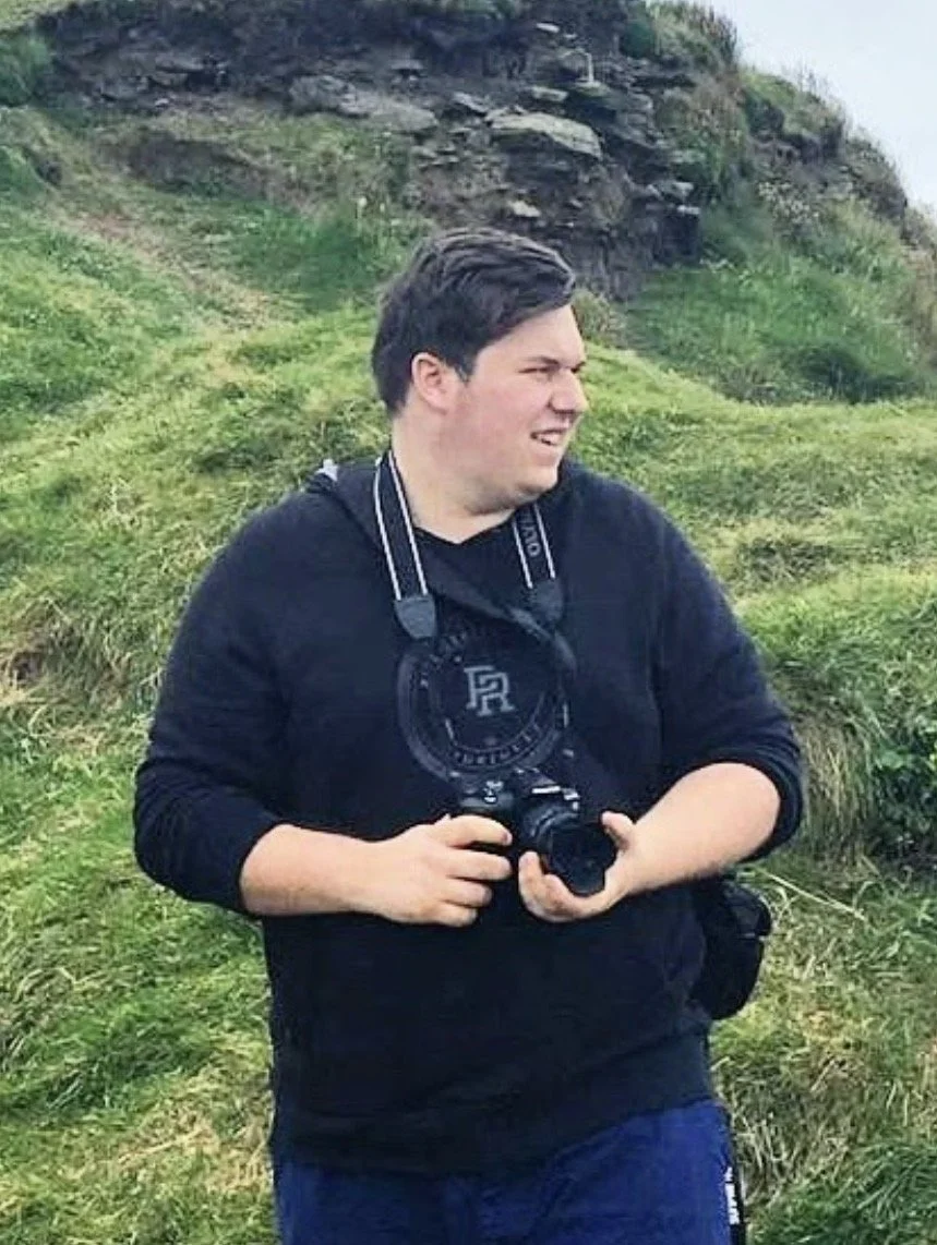 A young man with dark hair and a black jacket holding a camera, standing outdoors on a grassy hillside with rocks and greenery in the background.
