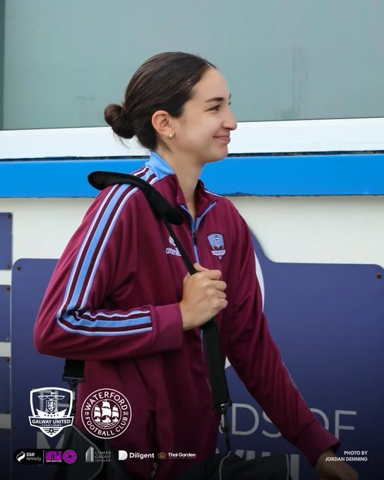 A young woman in a maroon sports jacket with a logo, carrying a black sports bag over her shoulder, standing outdoors near a blue and gray background.