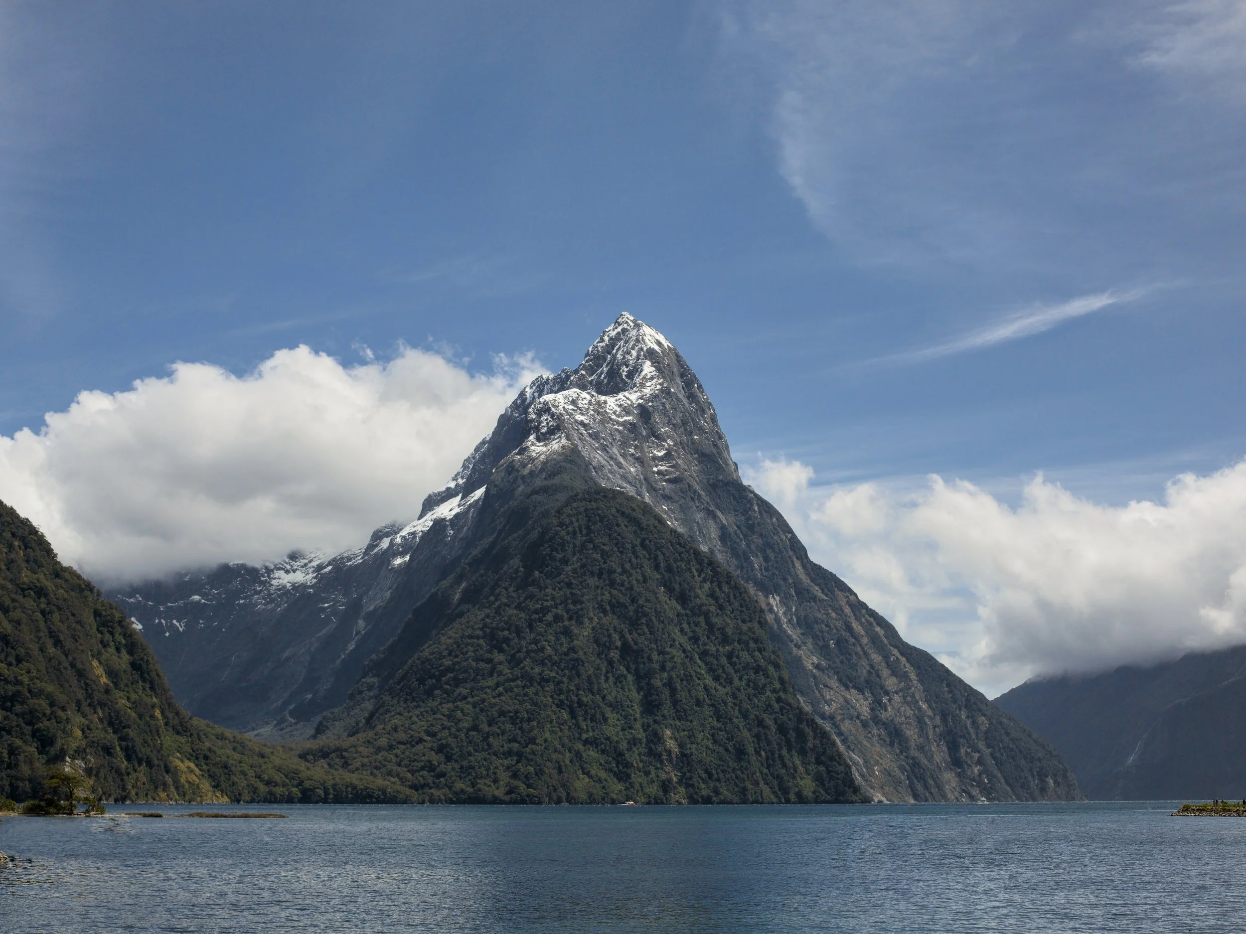 A scenic view of a snow-capped mountain surrounded by green forests and a body of water in the foreground, under a partly cloudy blue sky.